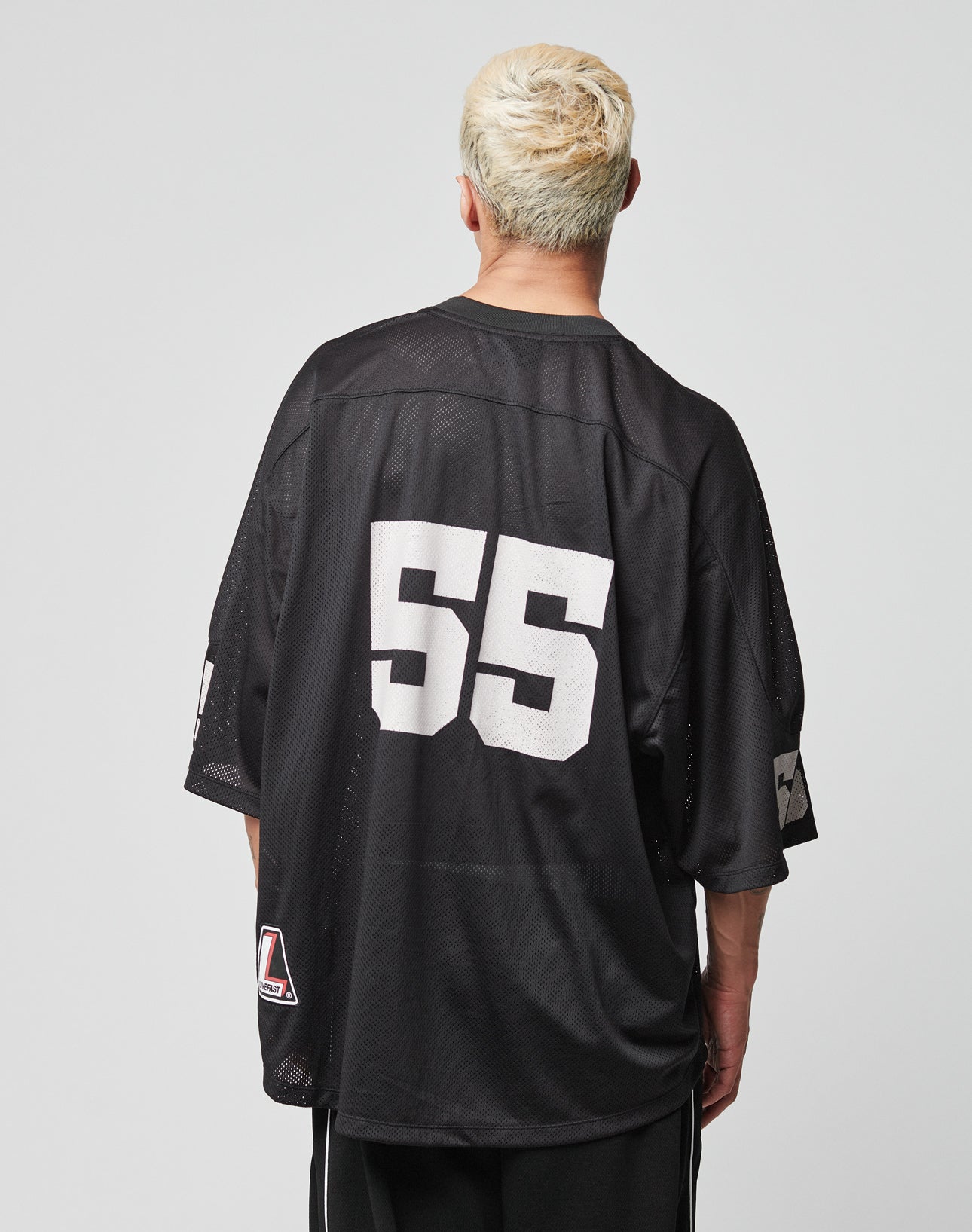 A person with short, bleached blonde hair is seen from behind wearing a black LIVE FAST DIE YOUNG LFDY Mesh Jersey with the number 55 in white, a V-Ausschnitt, and an oversize fit. The background is plain and light-colored.