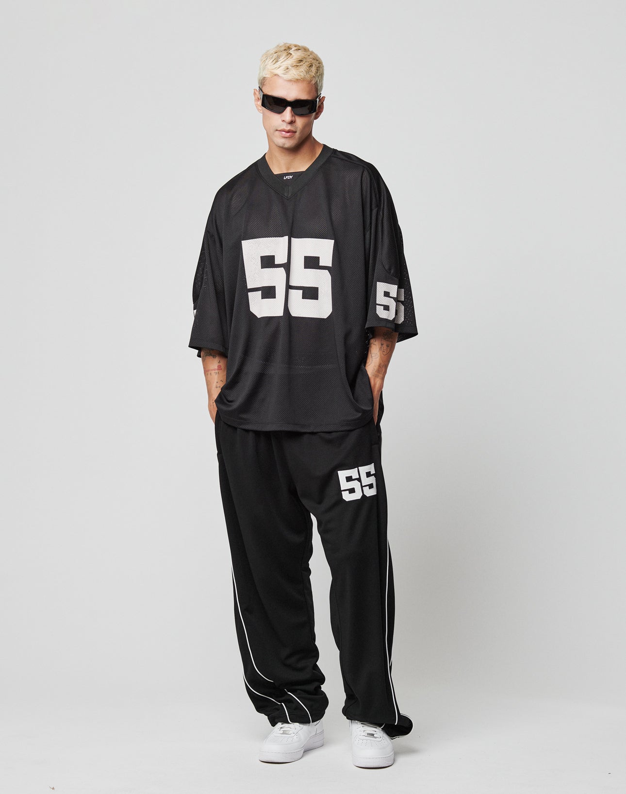 A person with short blond hair and sunglasses models LIVE FAST DIE YOUNG’s LFDY 55 Trackpants, loose black polyester track pants with an elastic waistband and white 55 detail, paired with a matching jersey and white sneakers against a plain background.