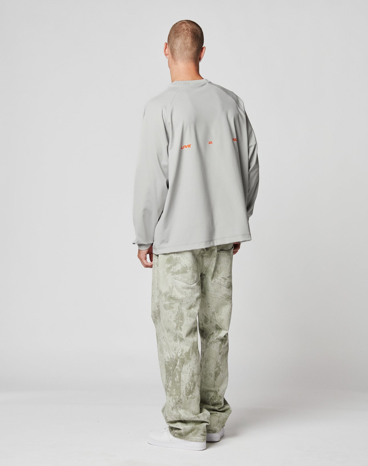 A person faces away from the camera, wearing a loose light gray LIVE FAST DIE YOUNG LFDY Sports Longsleeve with oversize fit, paired with light green patterned wide-leg pants and white shoes, set against a plain white background.