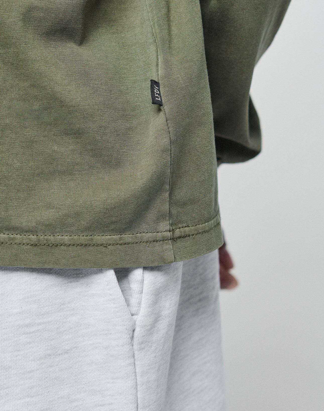 Close-up of a person wearing the LIVE FAST DIE YOUNG Basic Longsleeve in light gray (100% Baumwolle) with a pocket, olive green oversize fit, and a small black side-seam tag. Background is plain and light-colored.