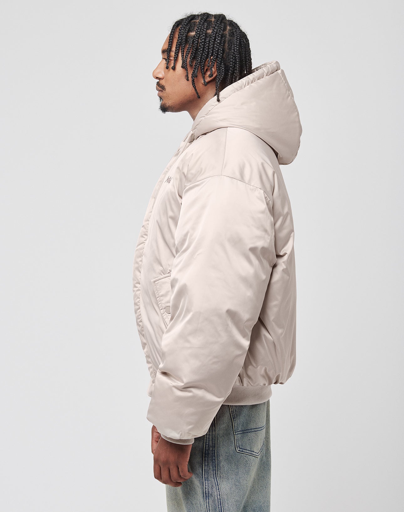 A man with braided hair, in profile, wears the LIVE FAST DIE YOUNG LFDY Winter Bomber Jacket in beige with hood and oversize fit, paired with light blue jeans, standing against a plain light gray background.