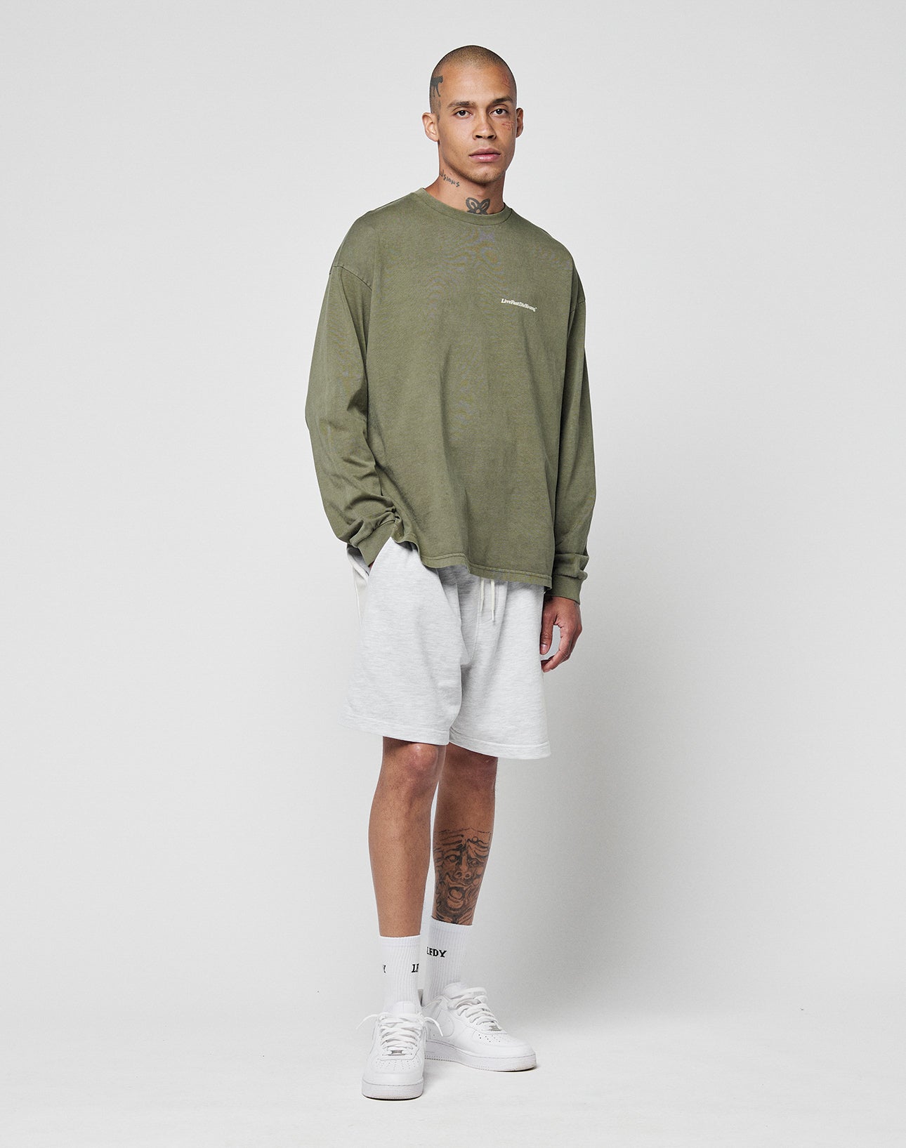 A person stands against a plain background wearing the LIVE FAST DIE YOUNG Basic Longsleeve in olive green (oversize fit, 100% Baumwolle), light gray shorts, white crew socks, and white sneakers. Tattoos are visible on their neck and left leg.