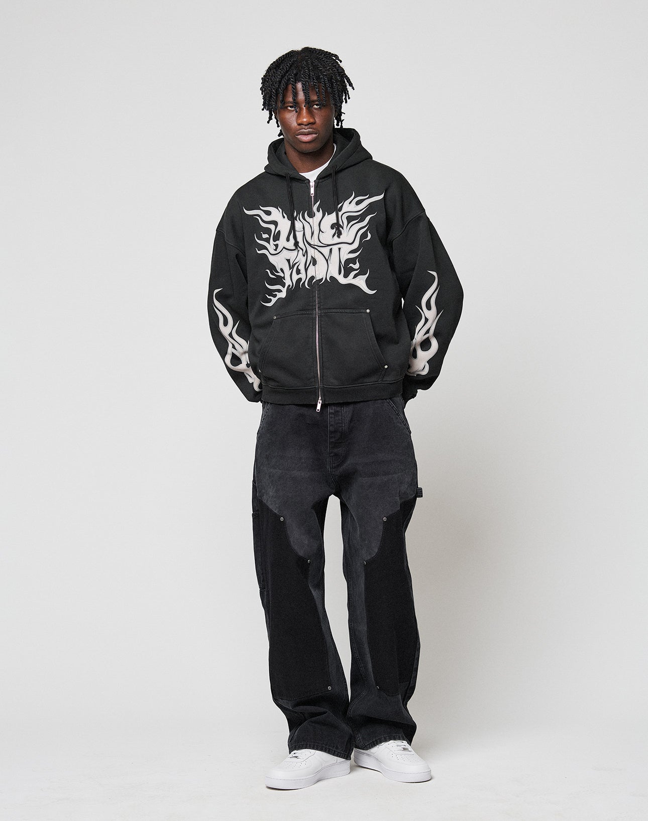A person with short dreadlocks wears the LIVE FAST DIE YOUNG LFDY Inferno Hooded Zip with white flame front and sleeve print, paired with black baggy jeans, white sneakers, and stands against a plain white background.