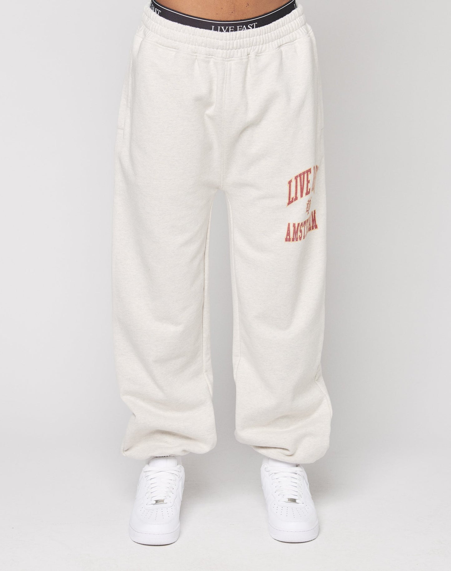 A person wears the LIVE FAST DIE YOUNG Live Fast Amsterdam Loose Jogger: cream, loose-fit sweatpants with an elastic waistband and red LIVE FAST lettering on the left thigh, made of soft cotton. Styled with white sneakers, black waistband, on a plain white background.