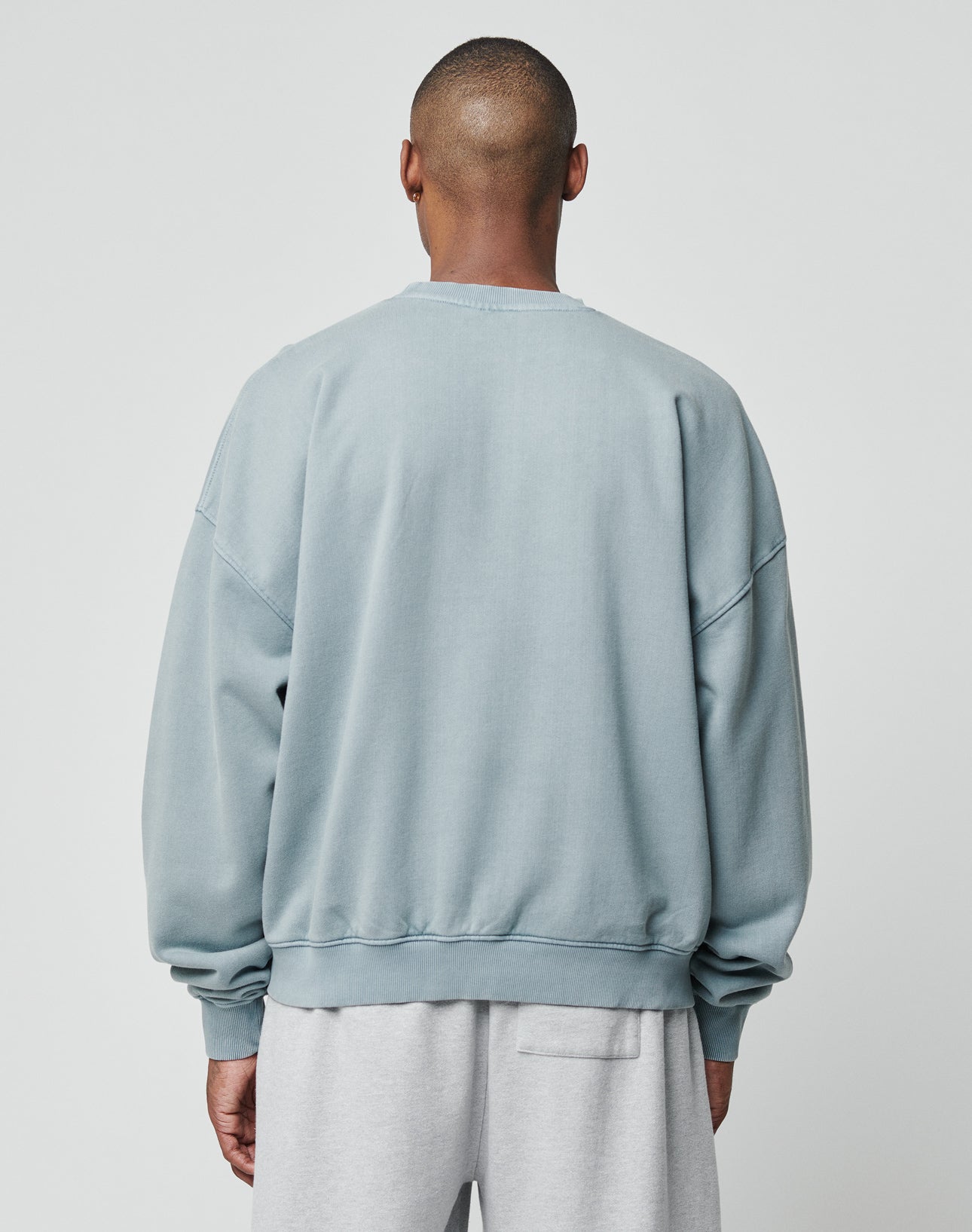 A person faces away from the camera wearing the LIVE FAST DIE YOUNG Ripped College Sweater in light blue with vintage wash and light gray sweatpants, standing against a plain light gray background.