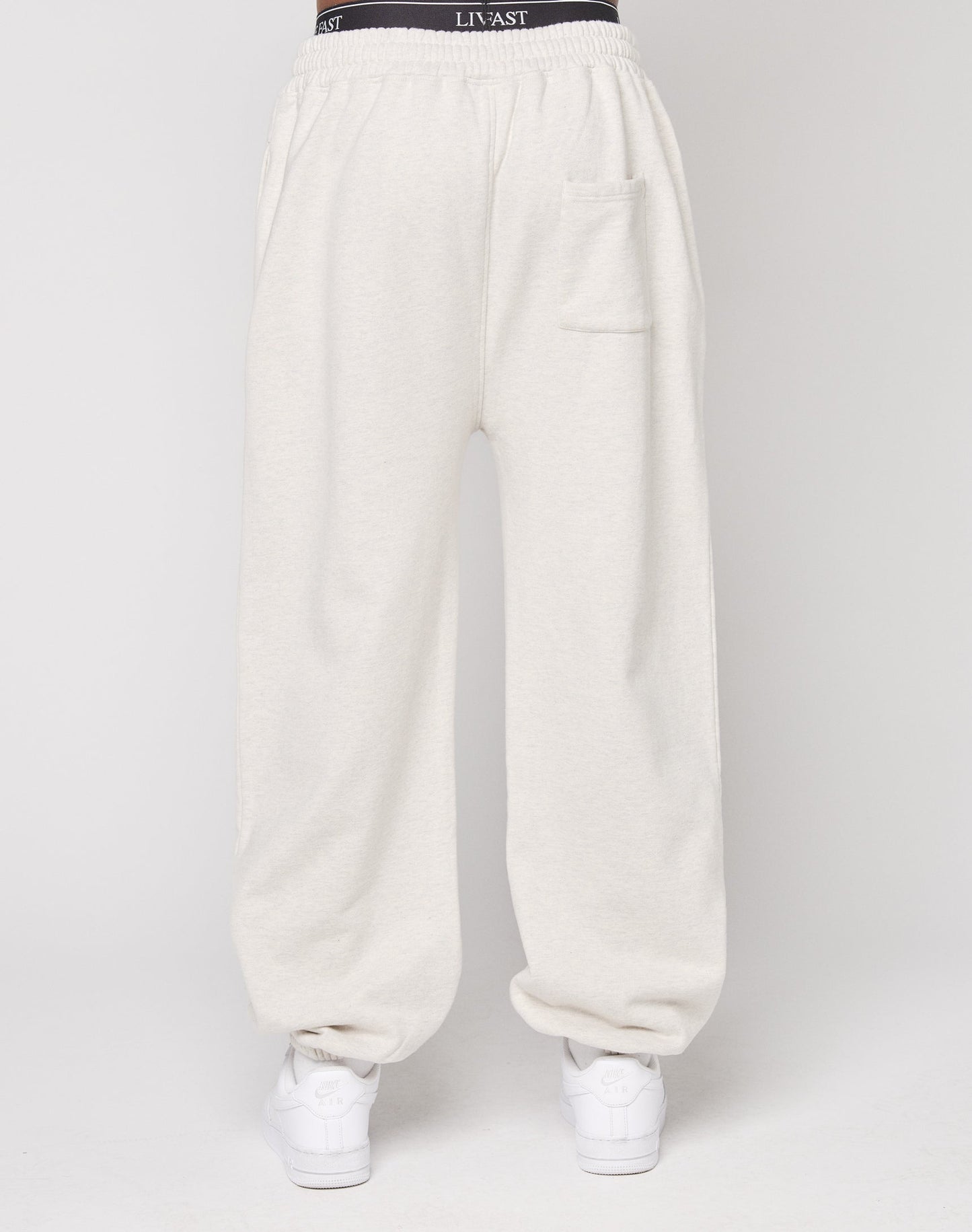 A person wears the Live Fast Amsterdam Loose Jogger by LIVE FAST DIE YOUNG—loose beige cotton sweatpants with a black elastic waistband featuring LIV & AST text, an oversize fit, and a right thigh front pocket, styled with white sneakers.