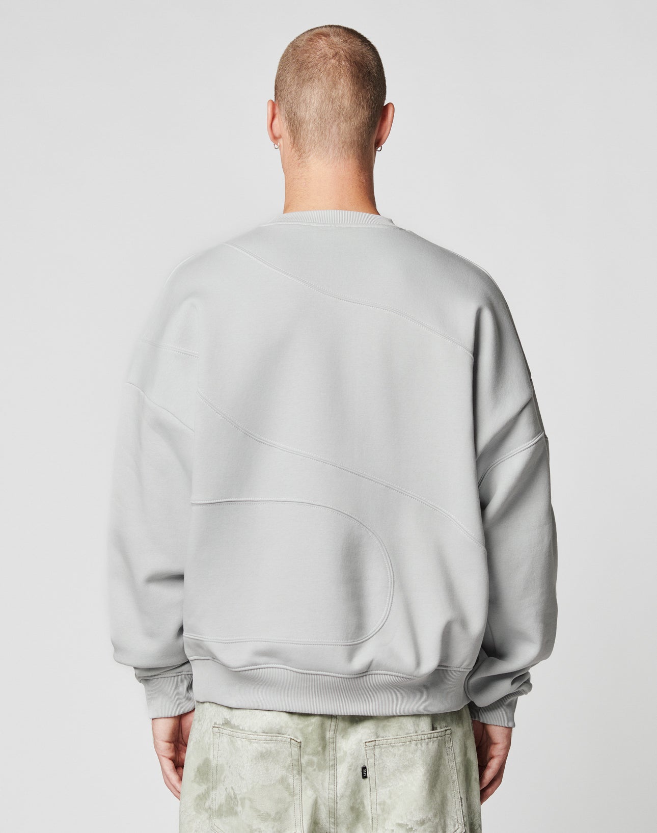 A person with a shaved head is seen from behind, wearing the LIVE FAST DIE YOUNG LFDY Wavy Sweater in light gray, featuring subtle curved stitching and an oversized fit, paired with textured light gray pants. The background is plain and light.