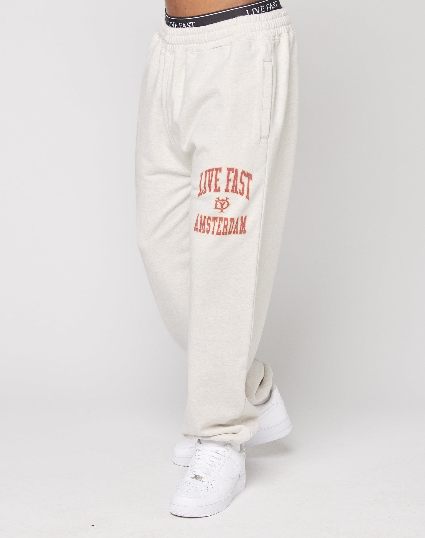 Model wears the LIVE FAST DIE YOUNG Live Fast Amsterdam Loose Jogger in off-white, featuring red LIVE FAST AMSTERDAM lettering on the left thigh, elastic waistband, and oversize fit. Styled with white sneakers; only lower body is visible.