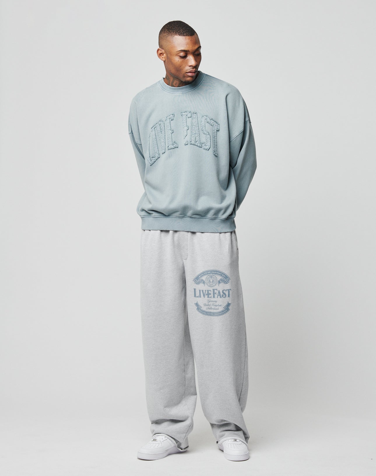 A man stands against a plain background wearing the Ripped College Sweater from LIVE FAST DIE YOUNG in light blue with a distressed print and matching oversize gray sweatpants, creating a relaxed look. He gazes down and to the side.