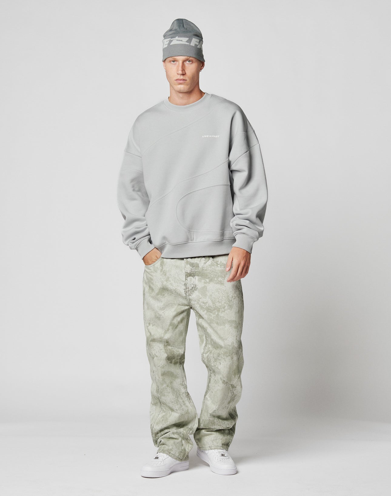 A man stands in a studio wearing an oversize fit light gray beanie, the LFDY Wavy Sweater by LIVE FAST DIE YOUNG, light greenish-gray patterned jeans, and white sneakers against a plain light background.