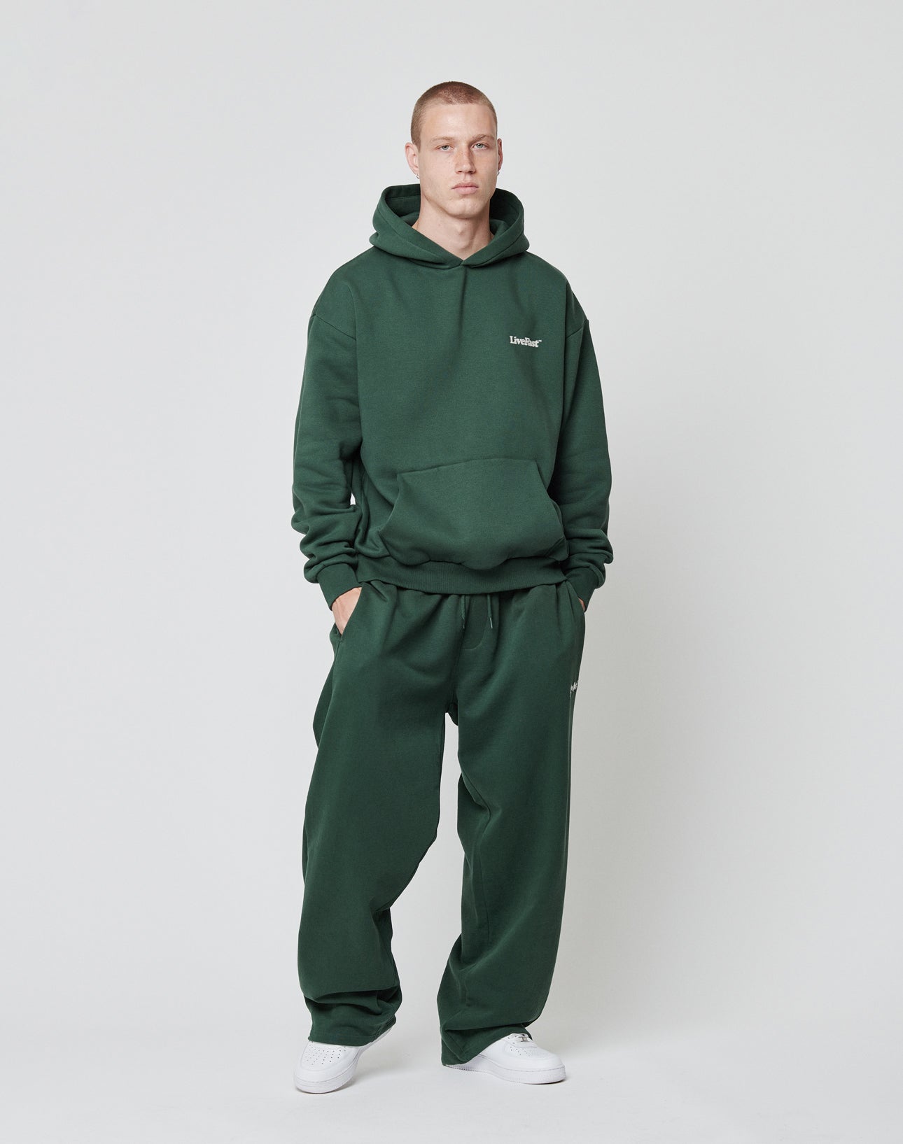 A man stands against a plain background wearing the LIVE FAST DIE YOUNG Basic 360 Hooded and matching sweatpants in dark green (70% Baumwolle) with front and back prints, hands in pockets, expression neutral.
