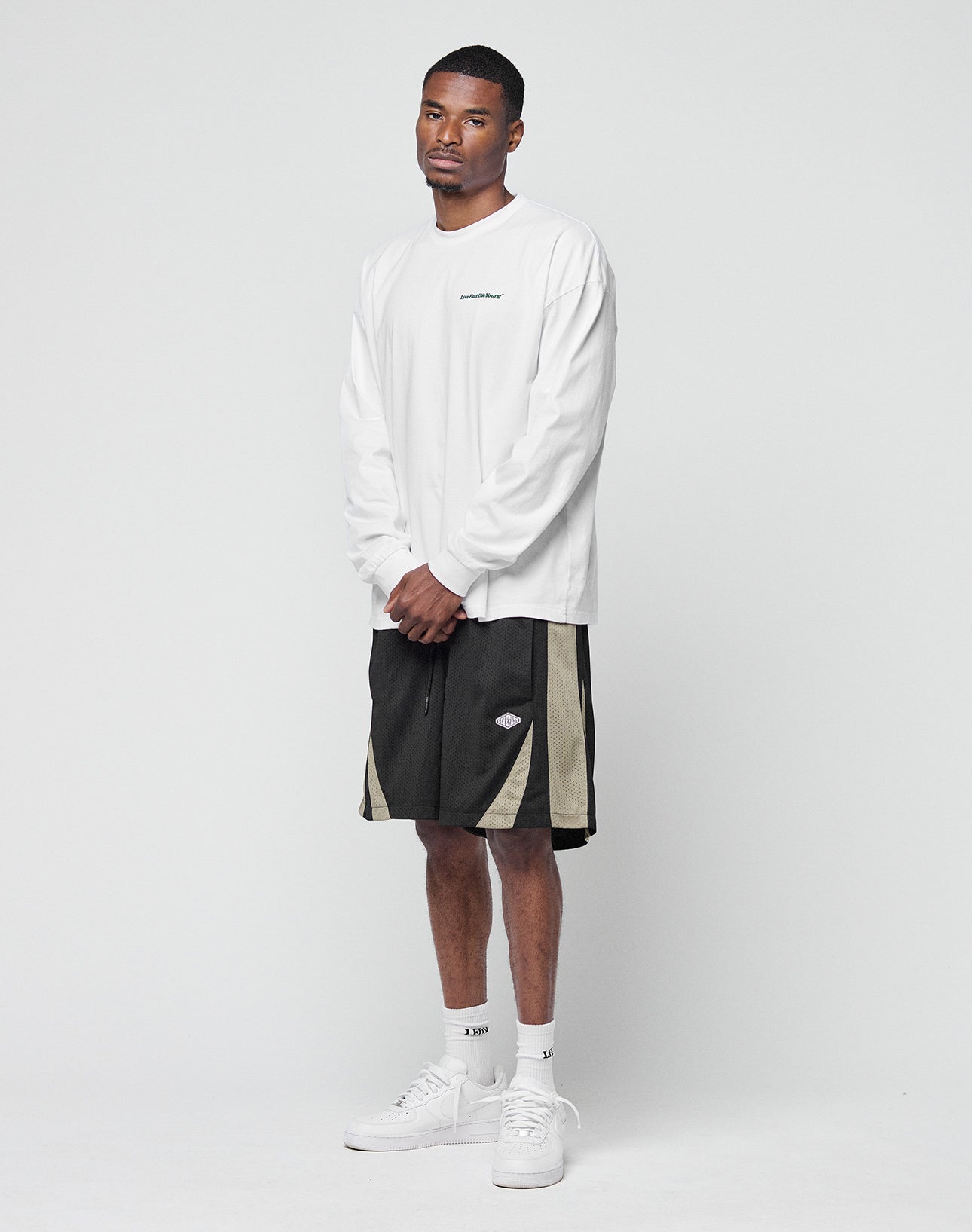 A man stands against a plain white background wearing a LIVE FAST DIE YOUNG Basic Longsleeve in white, black and beige athletic shorts, white crew socks, and white sneakers. His hands are clasped as he looks calmly at the camera.