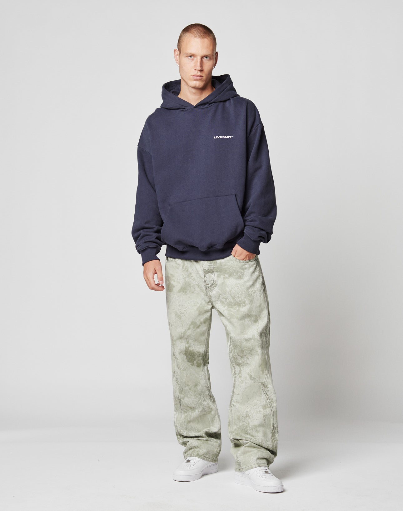 A man faces forward in an oversized navy Live Fast Plain Hooded sweatshirt by LIVE FAST DIE YOUNG, paired with light green tie-dye jeans and white sneakers, set against a plain light gray background.