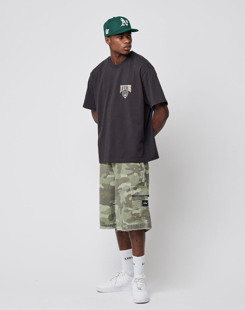 A man in a dark t-shirt, LIVE FAST DIE YOUNG Army Shorts with Cargotaschen, white sneakers, and a green baseball cap stands against a plain white background, gazing to the side with his hands in his pockets.