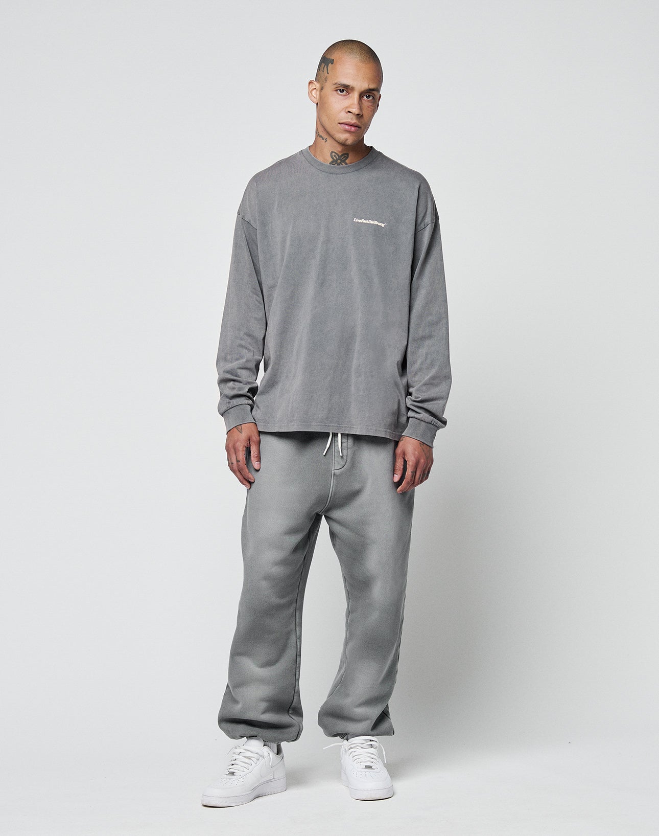 A person with short hair and visible neck and hand tattoos wears LIVE FAST DIE YOUNG’s Basic Loose Jogger set—gray, loose-fit, long-sleeve sweatshirt and sweatpants—paired with white sneakers. Subtle logo print details the outfit.