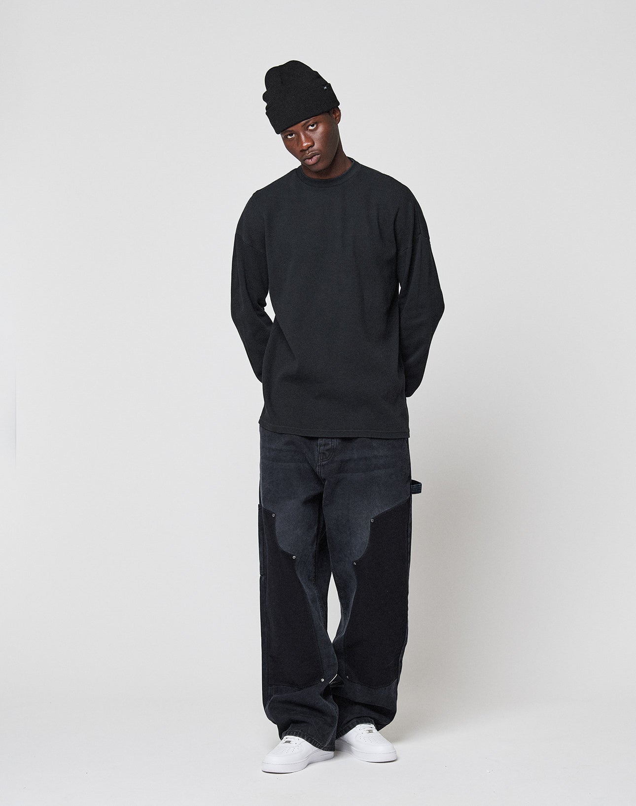 A person wears a LIVE FAST DIE YOUNG Blank Waffle Longsleeve in black, an oversize fit beanie, loose black patchwork pants, and white sneakers, standing against a plain light background with hands behind their back.