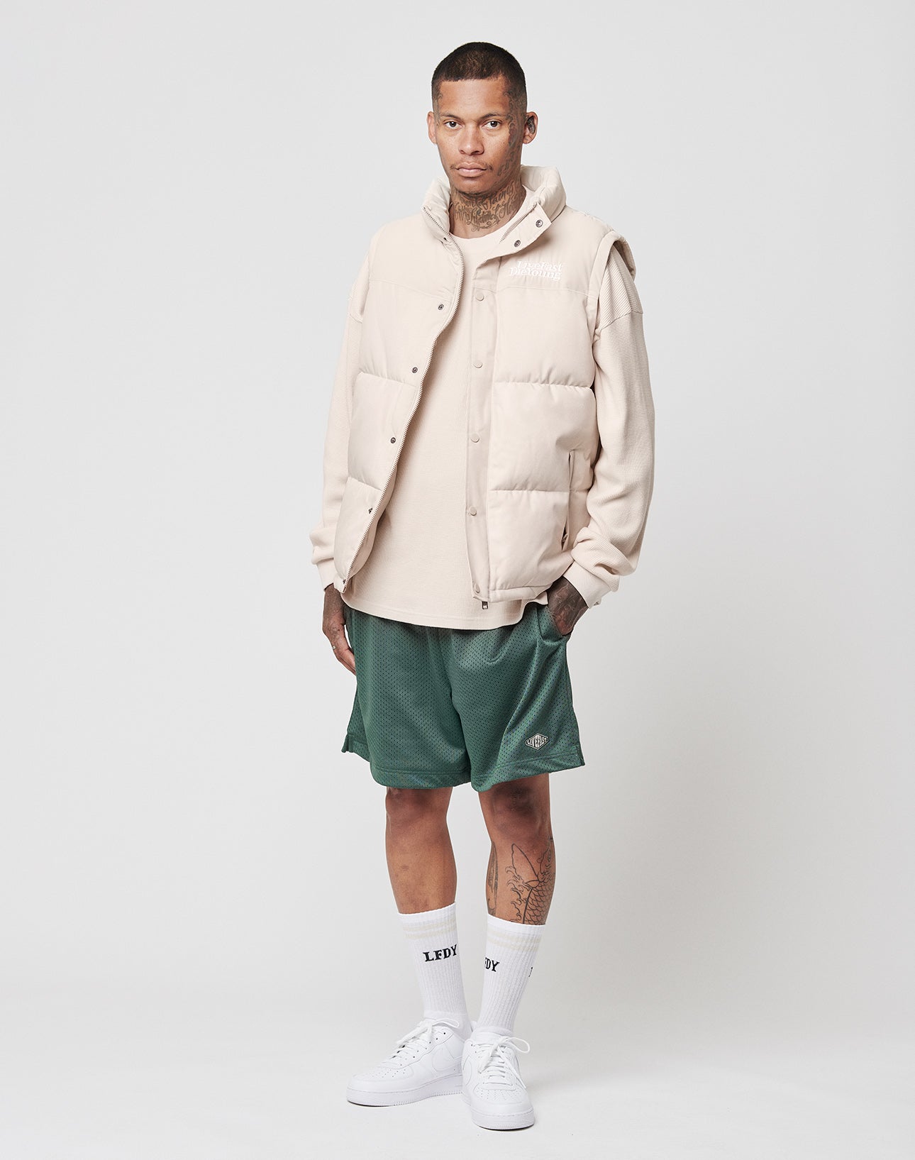 A person with arm tattoos and a neutral expression wears a beige polyester puffer vest, matching long-sleeve shirt, LIVE FAST DIE YOUNG Basic Mesh Shorts with elastic waistband in green, white LFDY socks, and white sneakers against a plain background.