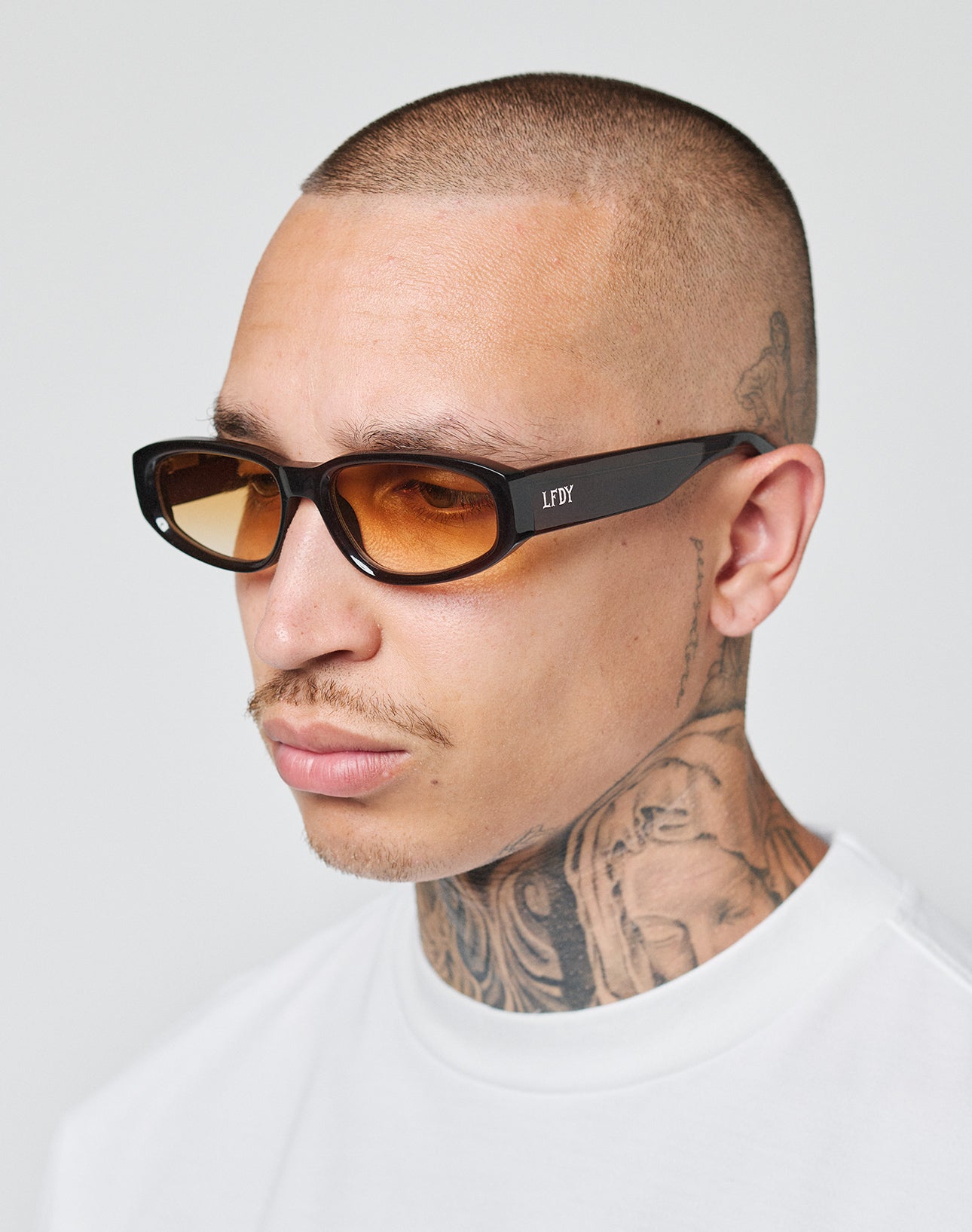 A man with a shaved head and tattooed neck wears LFDY x Chimi Shades 09 by LIVE FAST DIE YOUNG, featuring amber-tinted rectangular frames, paired with a white shirt against a plain light background.