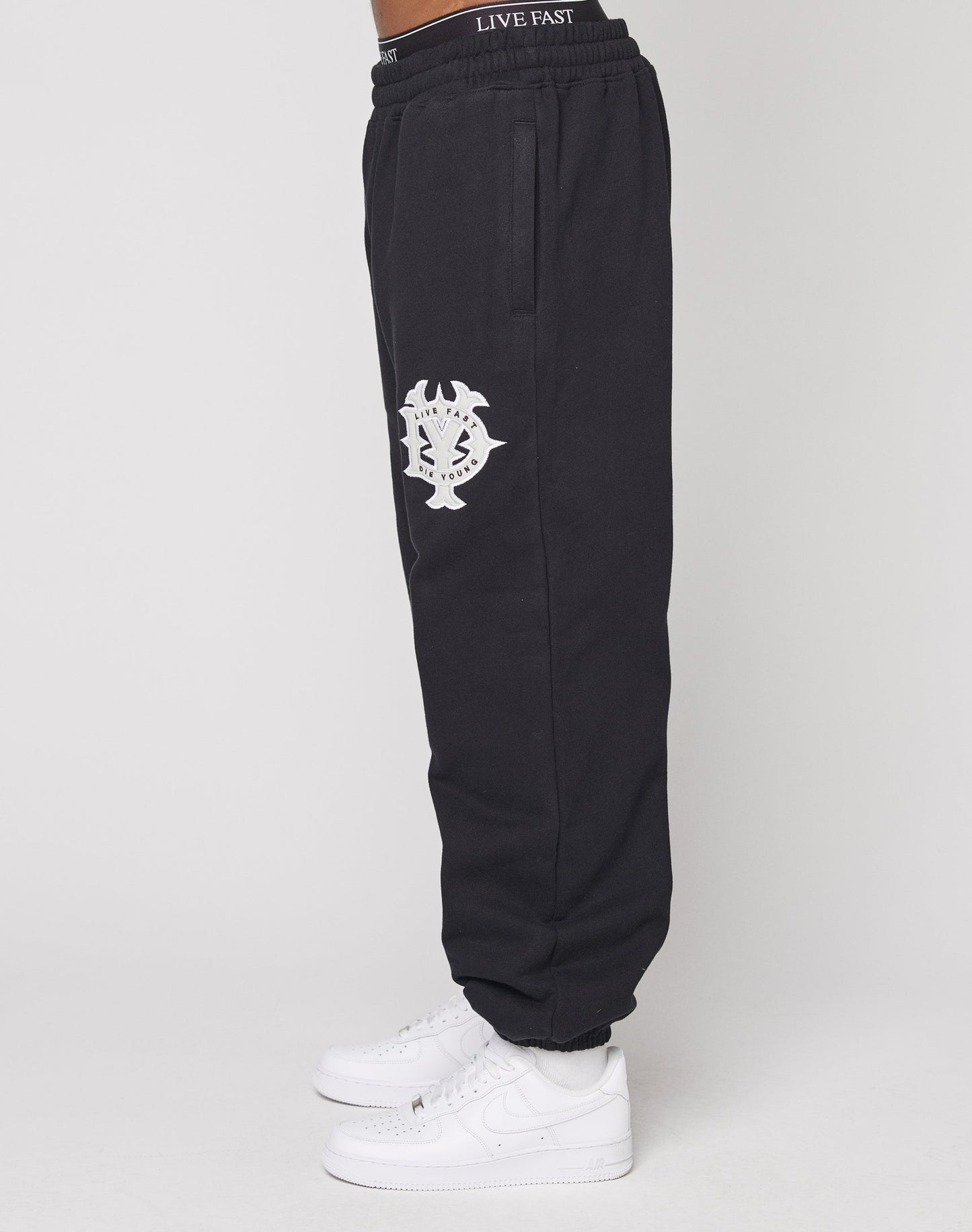 A person wearing DY Loose Jogger by LIVE FAST DIE YOUNG, featuring a white thigh graphic and an elastic waistband with "LIVE FAST," paired with white sneakers, standing sideways against a plain white background.