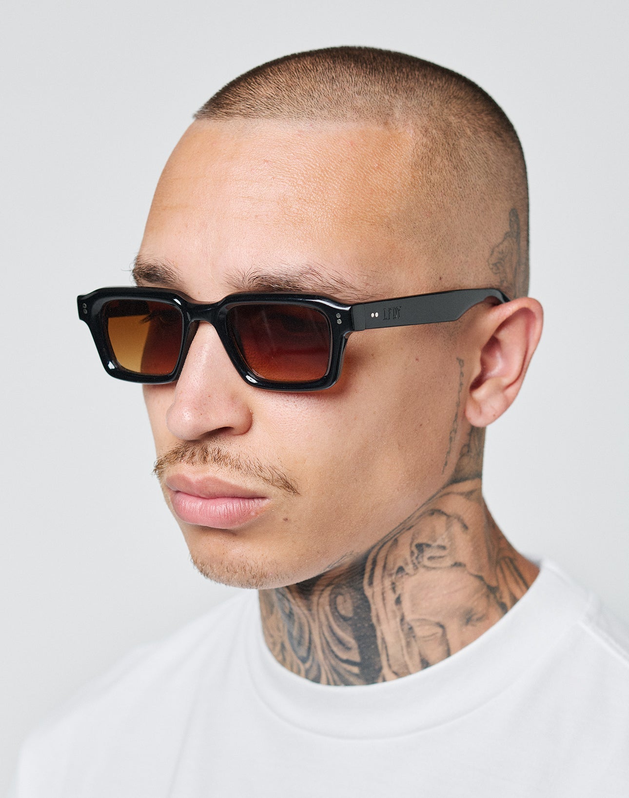 A man with a shaved head and neck tattoos wears LIVE FAST DIE YOUNG LFDY x Chimi Shades 03 and a white t-shirt, looking slightly to the side against a plain, light background.