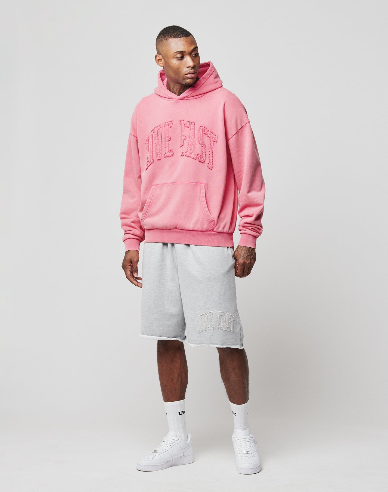 A man stands against a plain background wearing an oversize pink “LIVE FAST” hoodie, white socks, white sneakers, and light gray Ripped College Shorts by LIVE FAST DIE YOUNG. He looks slightly to his left.
