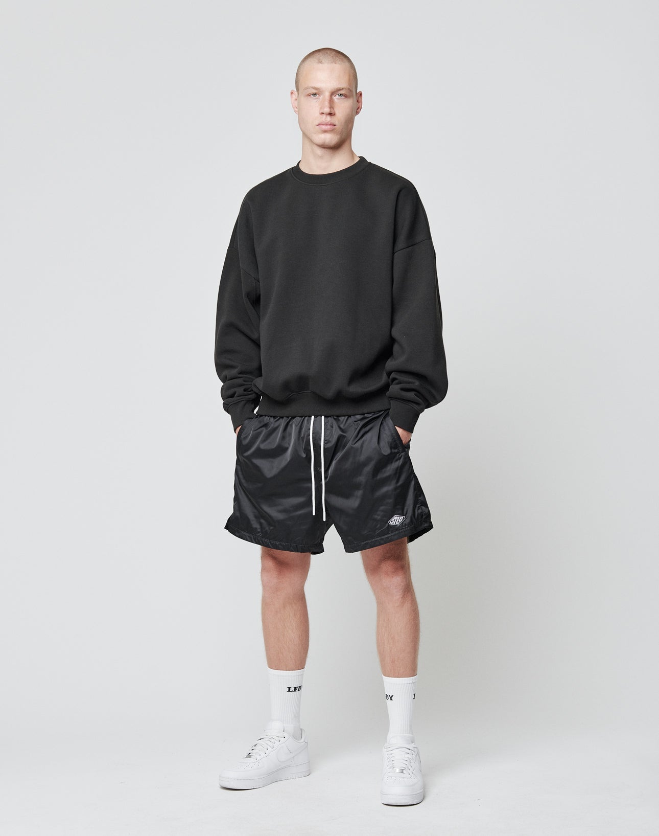 A man stands against a plain white background wearing a black sweatshirt, LIVE FAST DIE YOUNG Basic Satin Shorts in black with an elastic waistband and white drawstrings, plus white crew socks and sneakers. He faces the camera with a neutral expression.