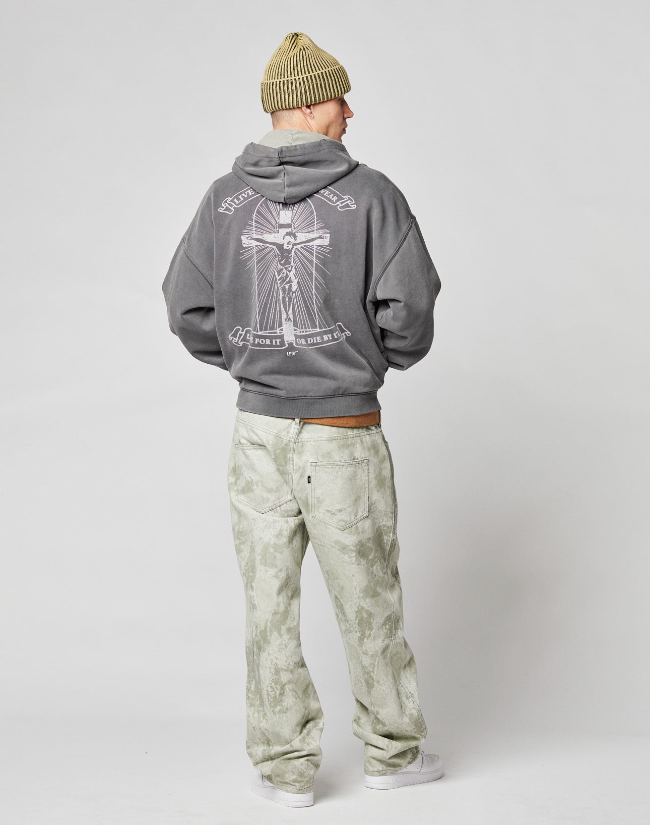 A person wearing the LIVE FAST DIE YOUNG LFDY Cross Hooded Zip, light greenish-gray pants, white sneakers, and a tan beanie stands facing away from the camera against a plain light background.