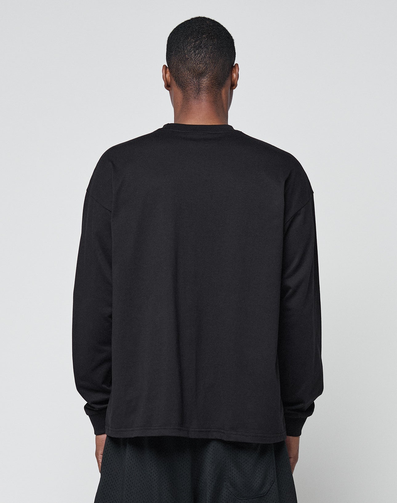 A person faces away from the camera, wearing LIVE FAST DIE YOUNGs Basic Longsleeve—an oversized black 100% Baumwolle shirt—and black shorts, all set against a light gray background.
