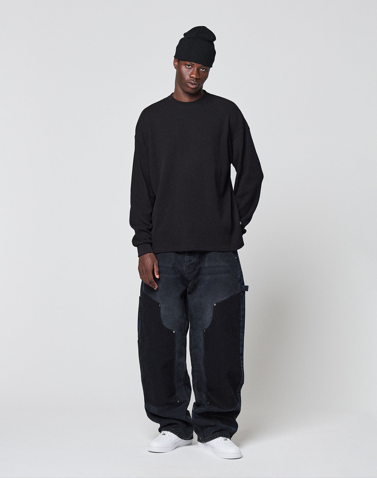 A person wears a black Blank Waffle Longsleeve by LIVE FAST DIE YOUNG with Ripp-Bündchen and Oversize Fit, loose dark jeans with light patches, and white sneakers, standing relaxed against a plain white background.