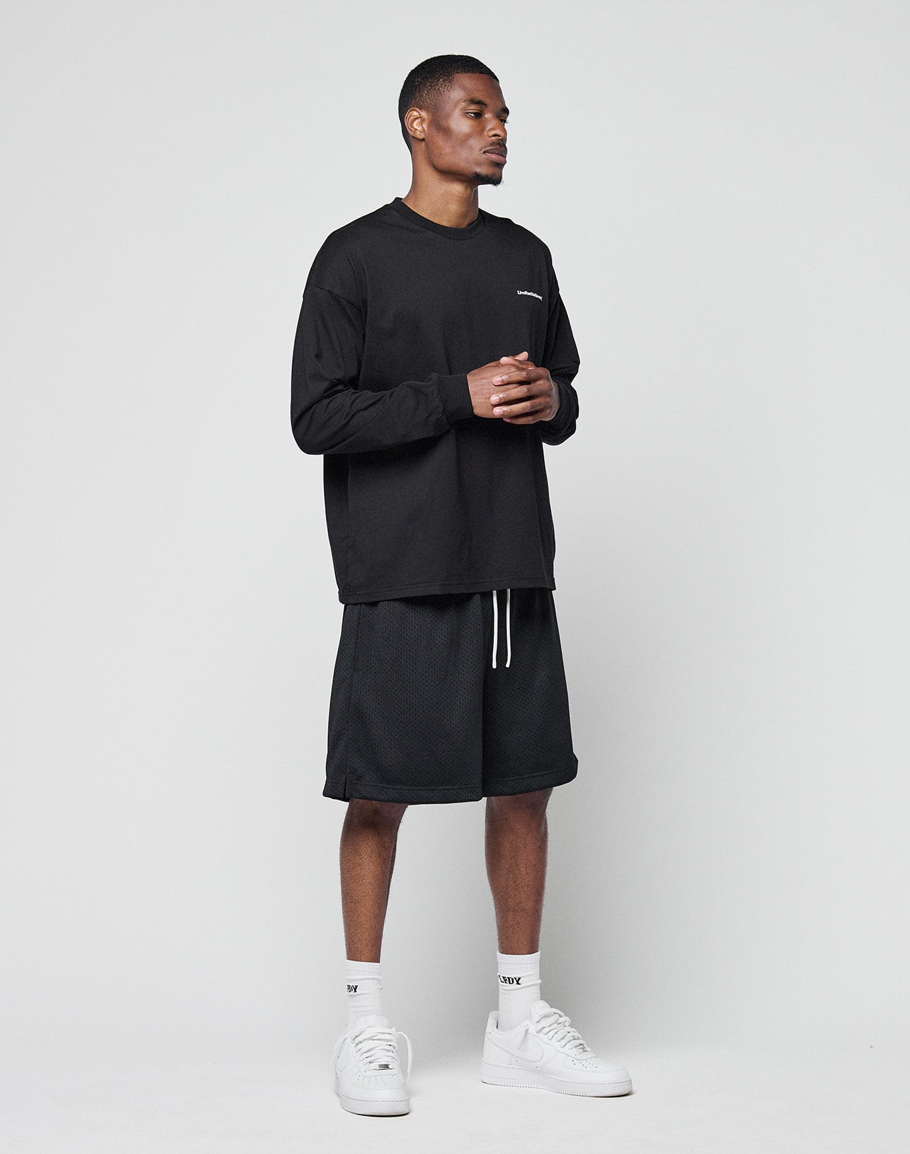A man stands against a plain light gray background wearing a black LIVE FAST DIE YOUNG Basic Longsleeve, black knee-length shorts, white socks, and white sneakers. He is facing slightly right with his hands clasped together.