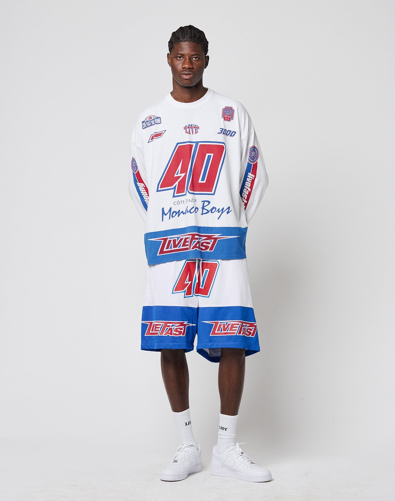 A man stands against a plain background wearing LIVE FAST DIE YOUNG’s LFDY Racing Mesh Shorts and a matching racing-style shirt, both featuring large prints, bold red, blue, and white graphics, numbers, and text. He pairs them with white sneakers and socks.