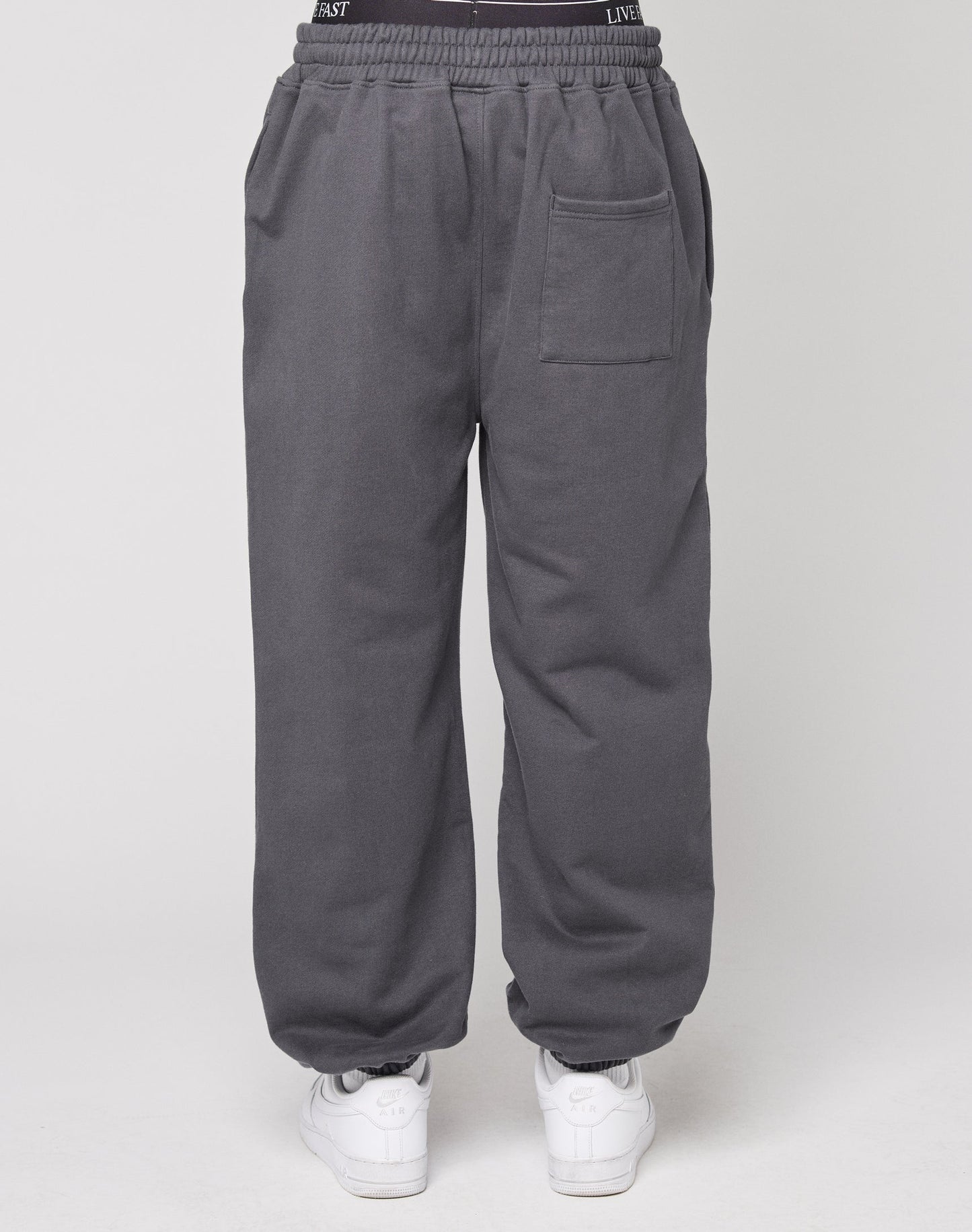Eine Person trägt den DY Loose Jogger von LIVE FAST DIE YOUNG in dunkelgrau mit Elastikbund und weißen Sneakers, von hinten vor hellem Hintergrund zu sehen.