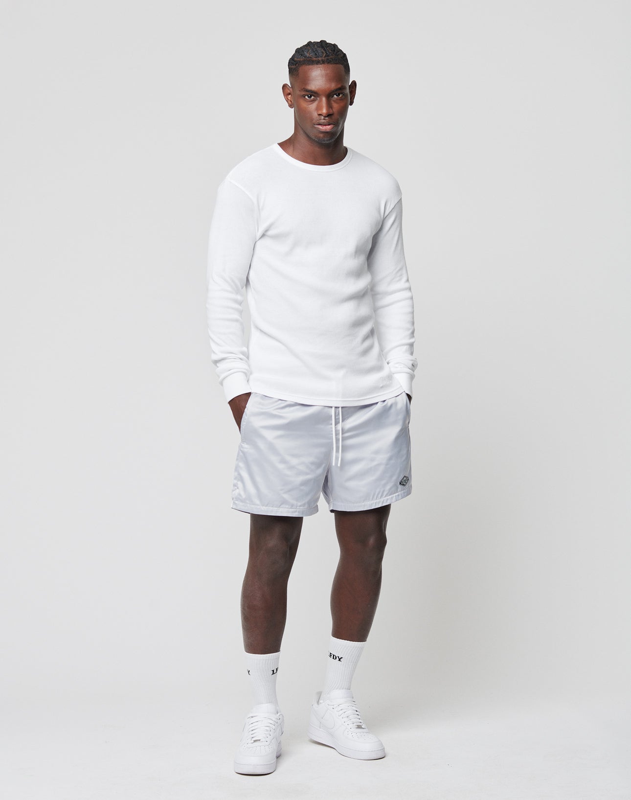 A man faces forward against a plain light background wearing a white long-sleeve shirt, LIVE FAST DIE YOUNG Basic Satin Shorts in light gray with a logo patch, plus white crew socks and sneakers. He has a neutral expression.