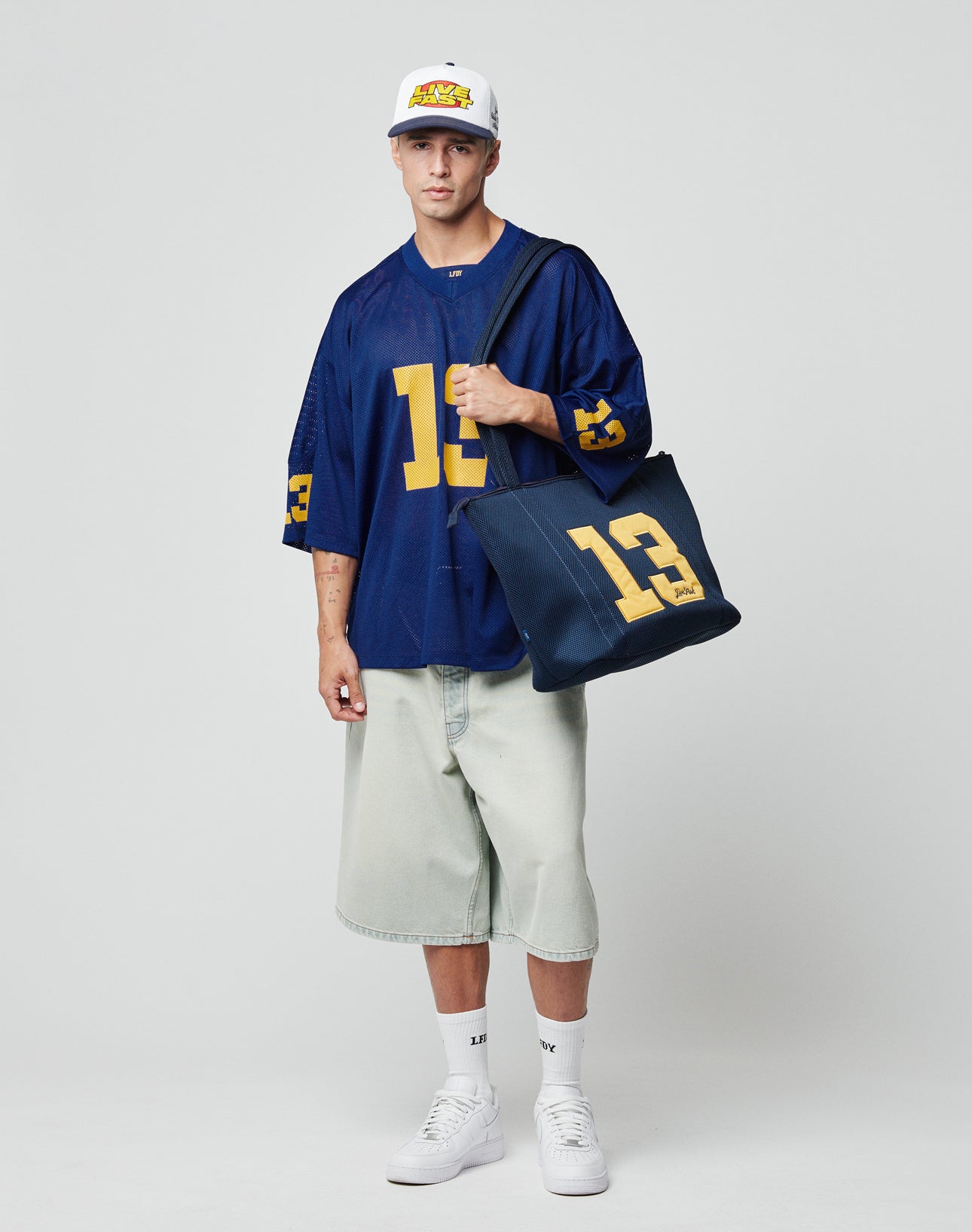 A person in a white cap, oversized blue “13” jersey, baggy denim shorts, and white sneakers holds the LIVE FAST DIE YOUNG LFDY Mesh Beach Bag with number 13 and Stick-Patch, standing against a plain light background.