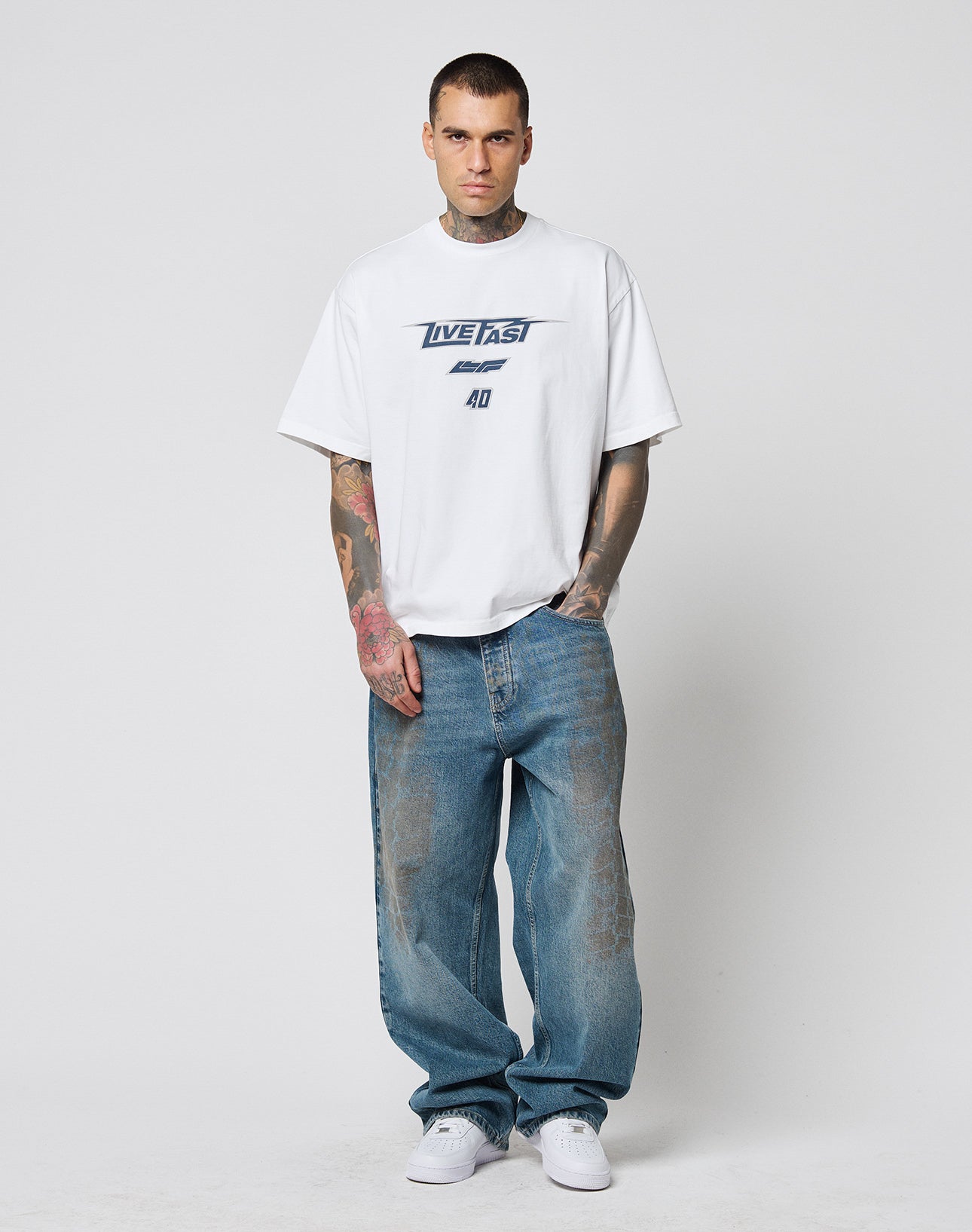 A man with short hair and tattoos wears the LFDY Triple Racing Tee by LIVE FAST DIE YOUNG, an oversized white T-shirt styled with baggy blue jeans and white sneakers, posing against a plain light background.
