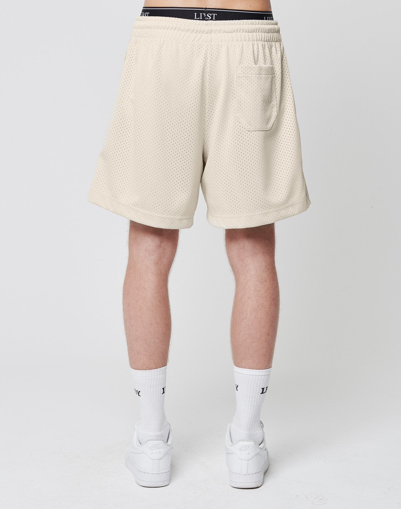 A person stands facing away, wearing LIVE FAST DIE YOUNG Basic Mesh Shorts in light beige with a regular fit, back pocket, and an LJST-branded waistband. They pair them with white socks and sneakers against a plain light-colored background.