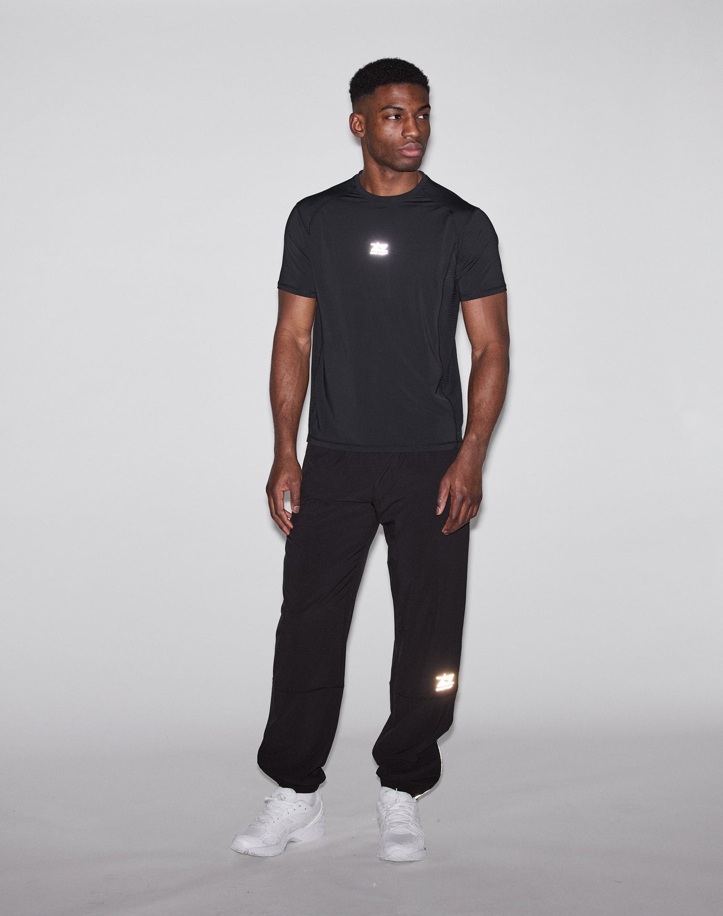 A man stands against a light gray background, wearing a black oversize athletic shirt and LIVE FAST DIE YOUNG Live Fast Performance Running Pants with reflective print and elastic waistband, plus white sneakers. He looks to his left, relaxed.