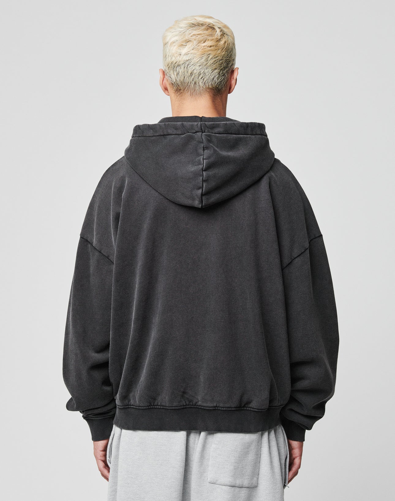 A person with short, bleached blonde hair is seen from behind wearing the LIVE FAST DIE YOUNG Ripped College Hooded Zip in Oversize Fit and light gray shorts. The 100% Baumwolle fabric contrasts with the plain, light-colored background.