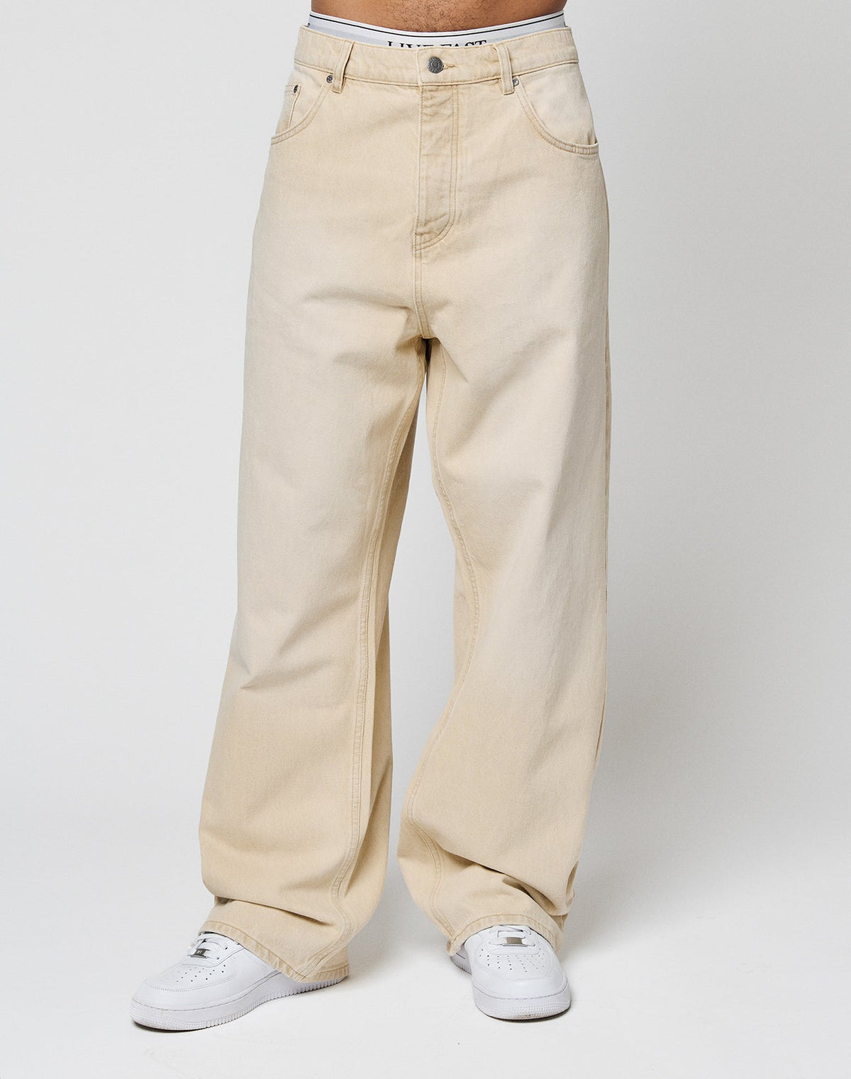 A person models the LIVE FAST DIE YOUNG LFDY Baggy Denim aus 100% Baumwolle in beige with white sneakers, standing against a plain white background. The cropped photo highlights the pants from waist to ankles.