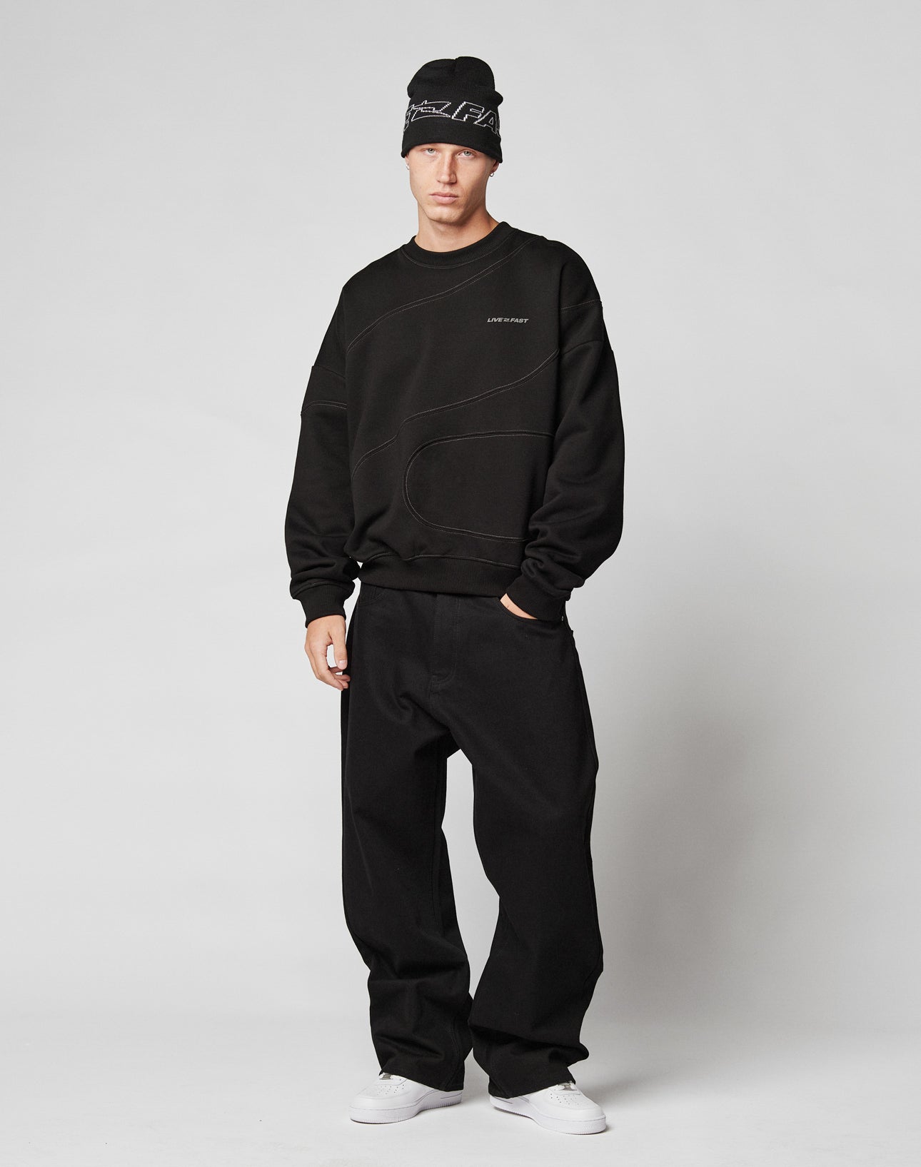 A person stands against a plain background wearing a black beanie, loose black pants, white sneakers, and the LFDY Wavy Sweater by LIVE FAST DIE YOUNG—an oversize fit sweatshirt in cotton with clear streetwear inspiration.