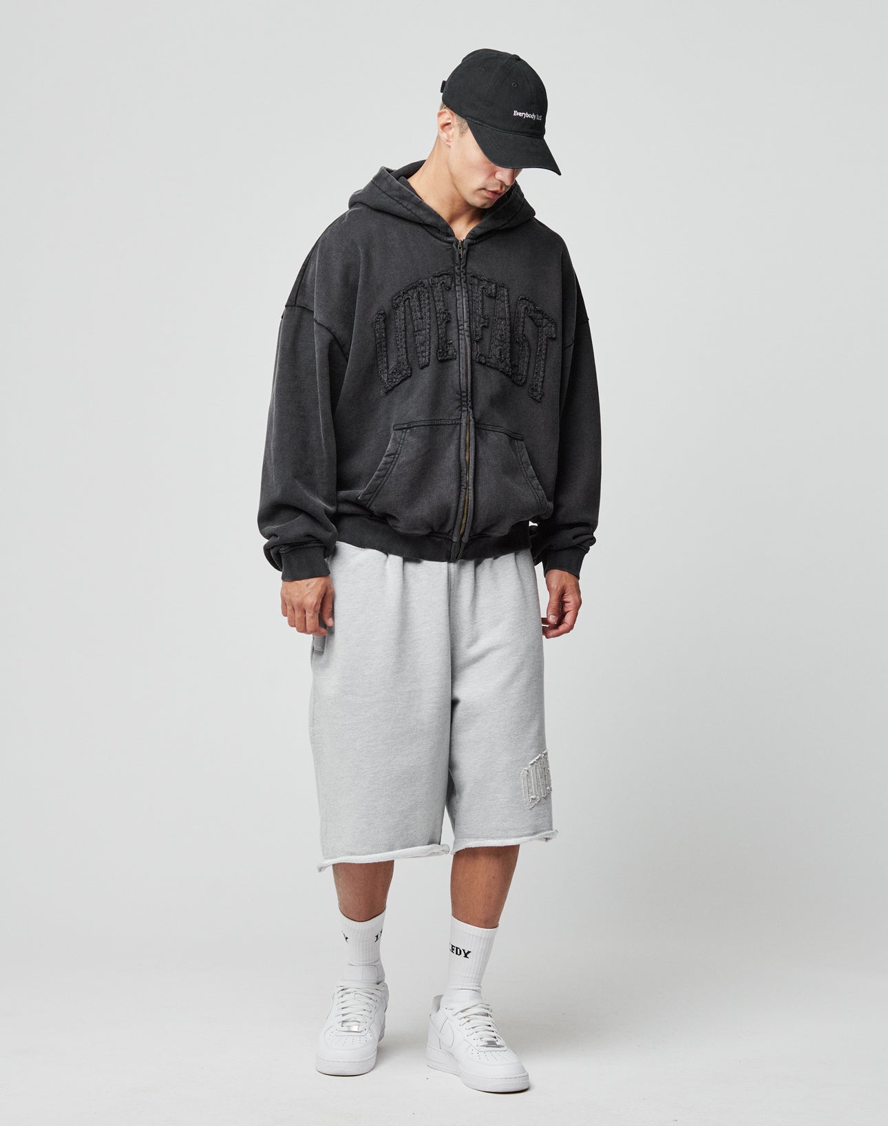 Wearing a black Ripped College Hooded Zip by LIVE FAST DIE YOUNG, vintage wash effect, loose light gray knee-length shorts, white socks, and white sneakers, a person stands against a plain light background.