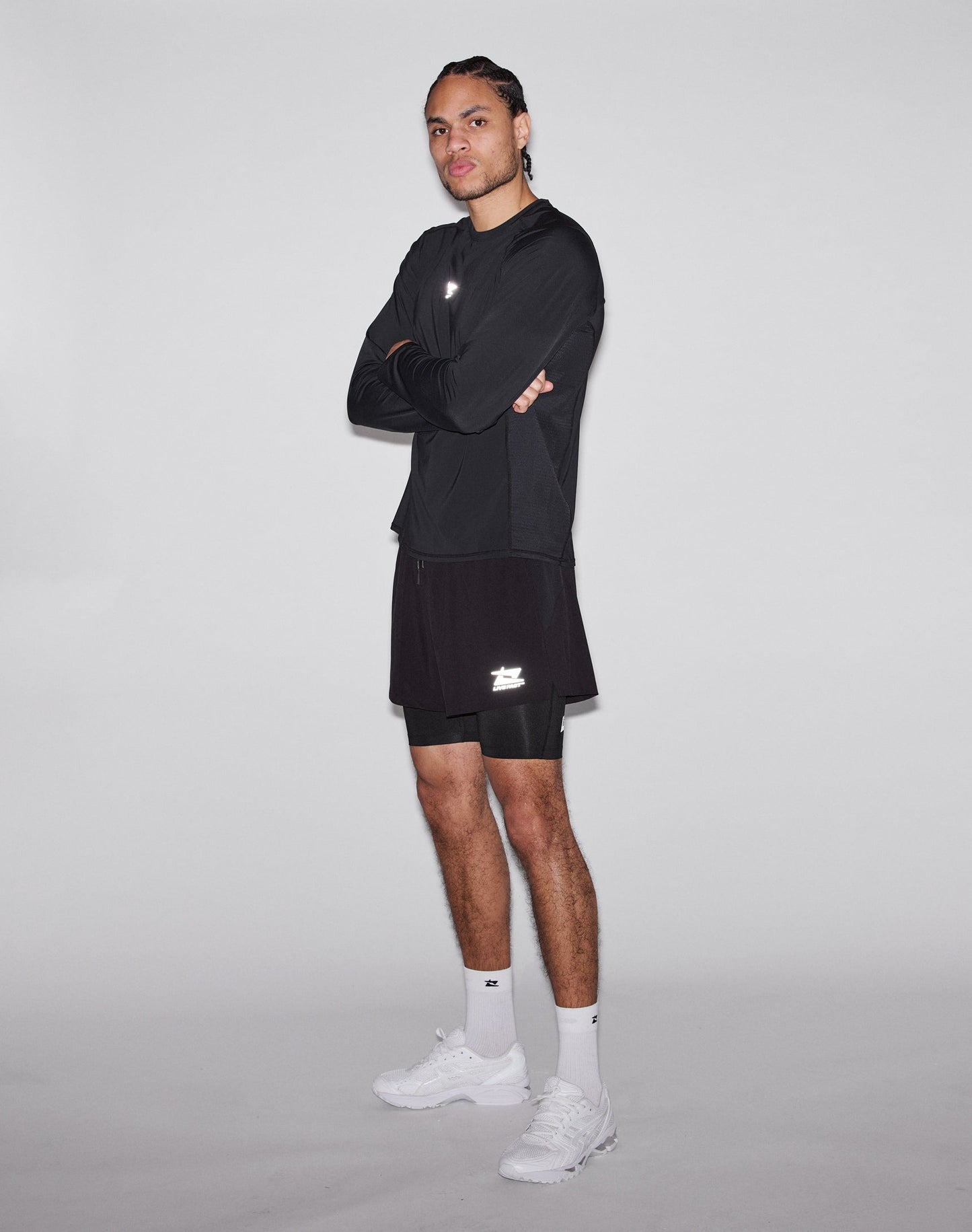 A person stands with arms crossed, wearing a black Regular Fit long-sleeve shirt, LIVE FAST DIE YOUNG Live Fast Performance Running Shorts in black with integrated tights, white socks, and white sneakers against a light-gray background.