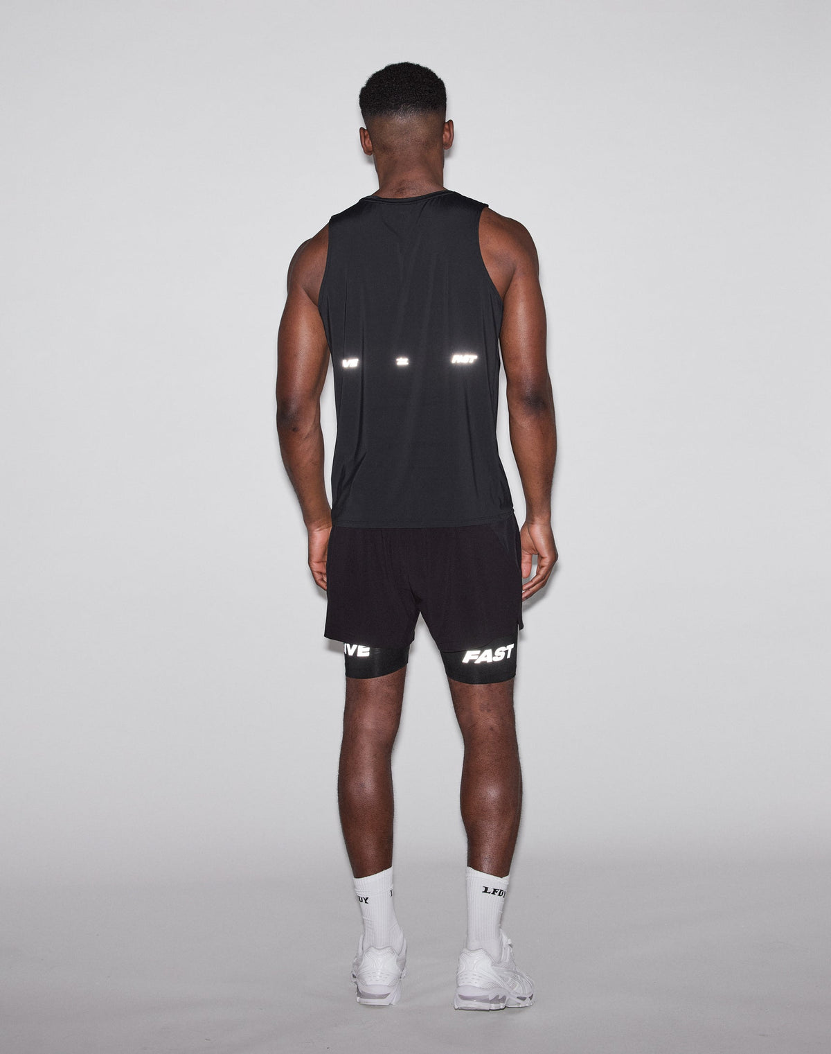 A man faces away from the camera, wearing a black LIVE FAST DIE YOUNG Live Fast Performance Tank, black shorts with FAST and LIVE in reflective print, white socks, and white sneakers against a plain light background.