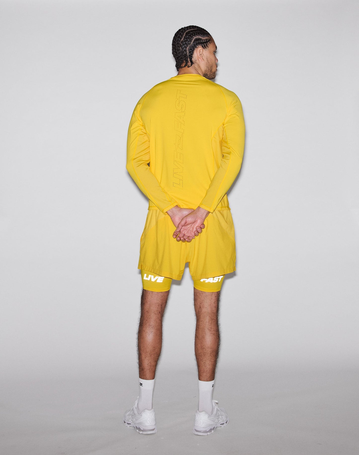 A person stands facing away, hands behind their back, wearing bright yellow Live Fast Performance Running Shorts by LIVE FAST DIE YOUNG with integrated tights and lightweight fabric, paired with a matching long-sleeve shirt and white sneakers against a light gray background.