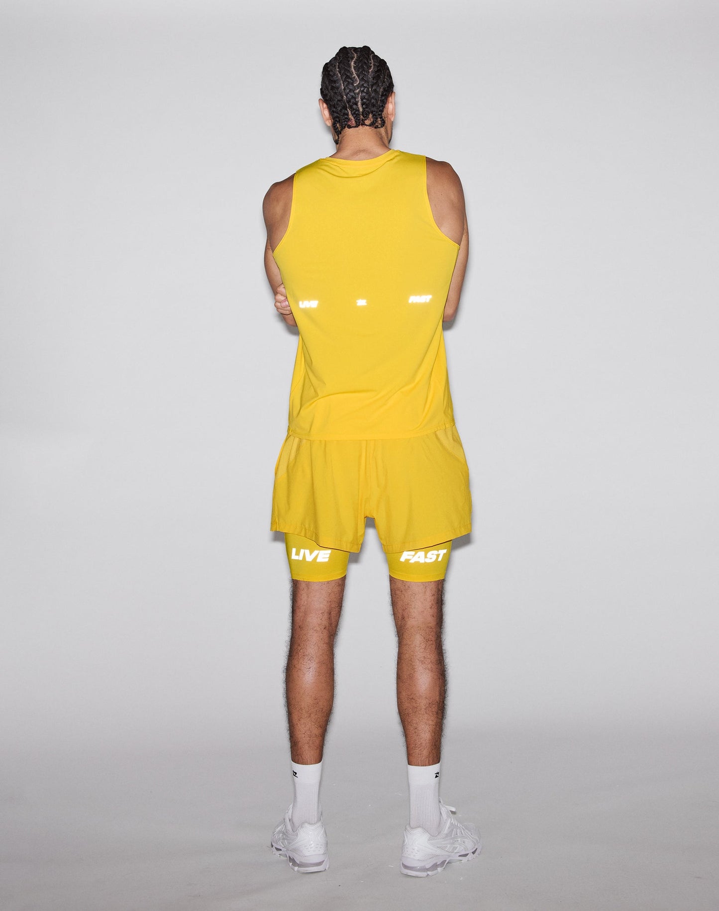 A person with braided hair wears the LIVE FAST DIE YOUNG Live Fast Performance Tank in yellow and matching shorts with reflective LIVE FAST print, plus white socks and sneakers. They face away from the camera with arms crossed against a plain background.