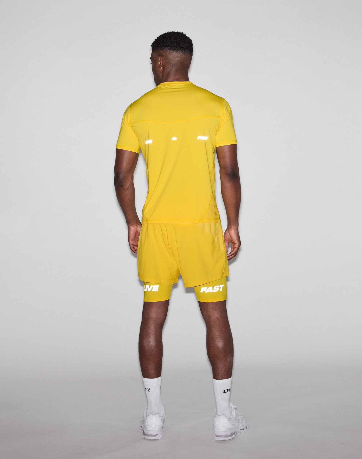 A man faces away from the camera in a slim-fit, bright yellow Live Fast Performance Tee by LIVE FAST DIE YOUNG, paired with matching shorts featuring LIVE and FAST, plus white socks and shoes. Reflective print details pop against a plain background.