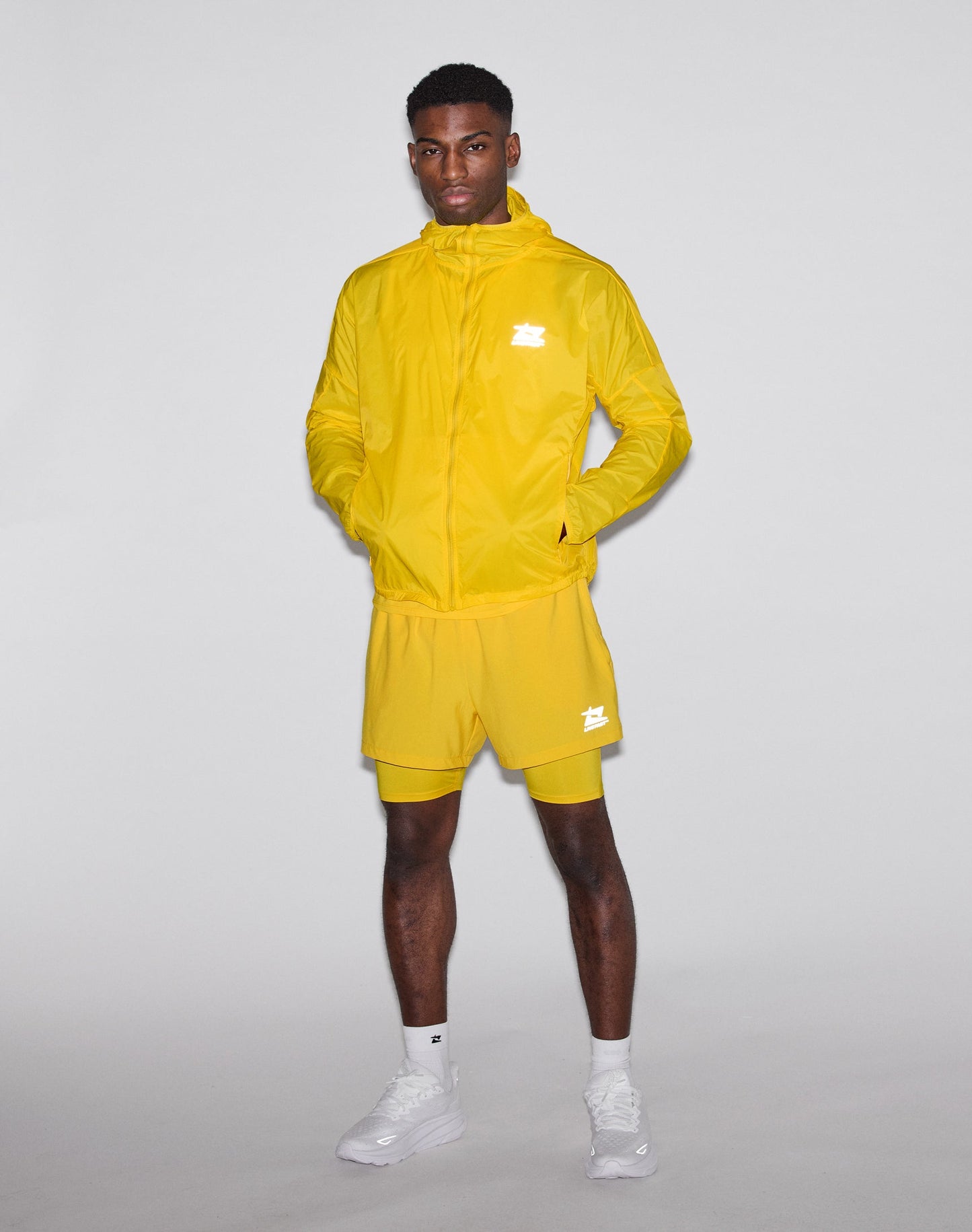 A man stands against a plain background wearing the bright yellow Live Fast Performance Running Jacket by LIVE FAST DIE YOUNG with reflektierender Frontprint, matching shorts, white socks, and white sneakers. Hands in pockets, he looks at the camera.