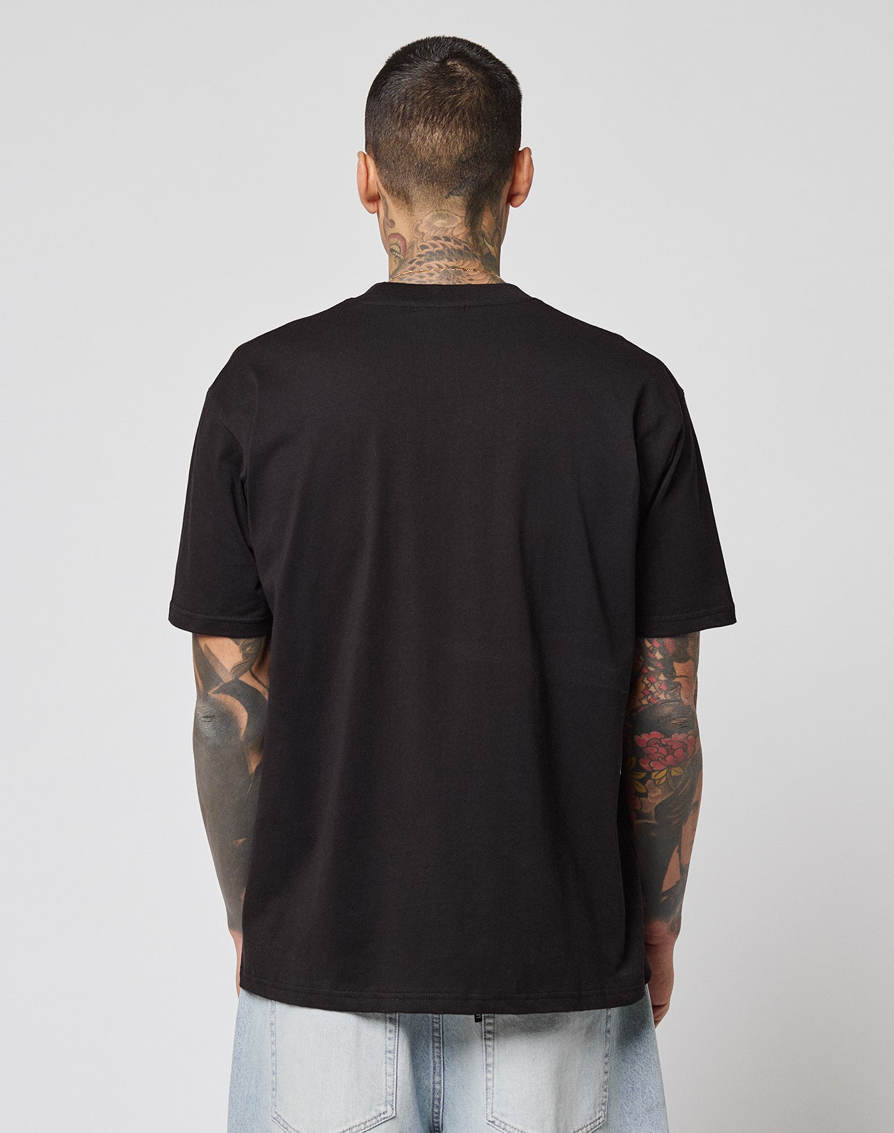 A person with short hair and tattoos, facing away, wears a LIVE FAST DIE YOUNG LFDY Sixers Tee in plain black and light blue jeans against a plain, light background.