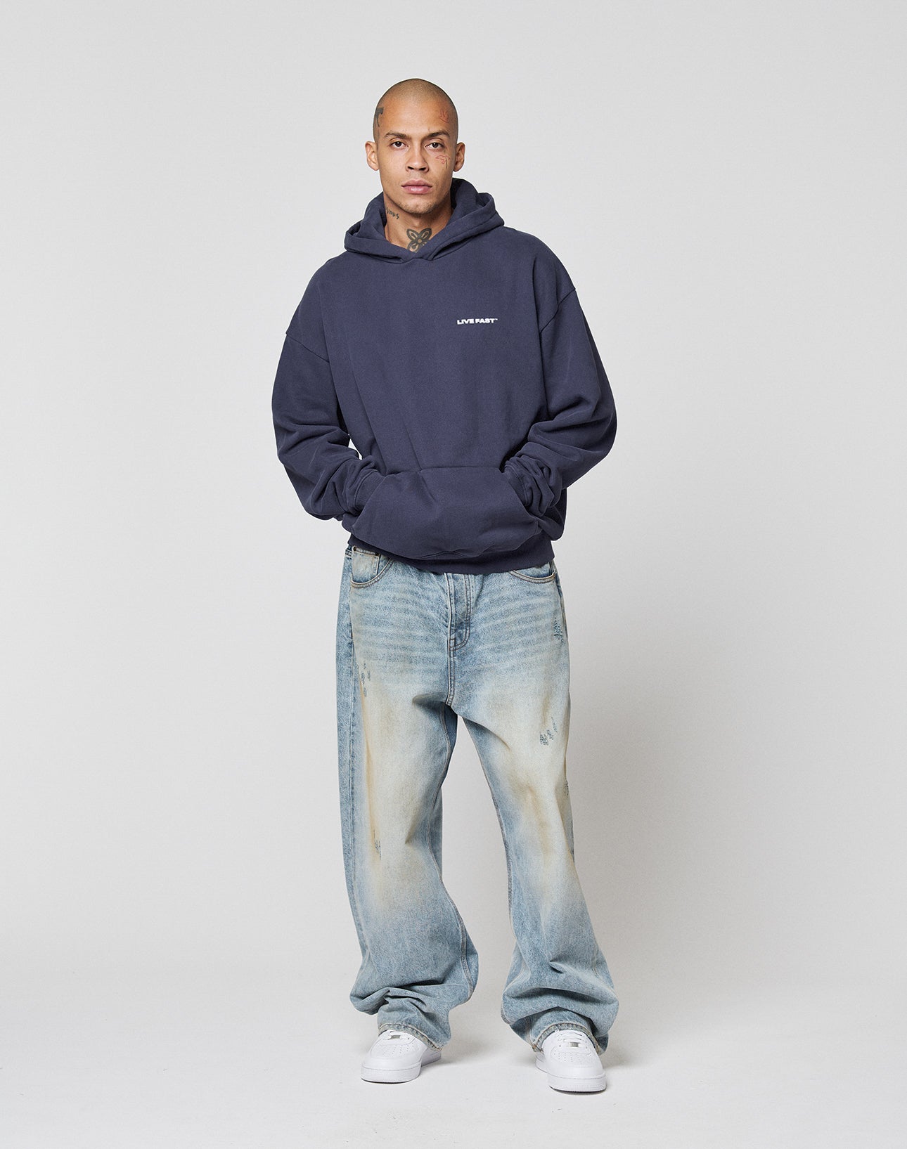 A person with a shaved, tattooed head stands against a light gray background, wearing the LIVE FAST DIE YOUNG Live Fast Plain Hooded navy blue hoodie with a bold front print, light blue baggy jeans, and white sneakers, hands in the hoodie pocket.