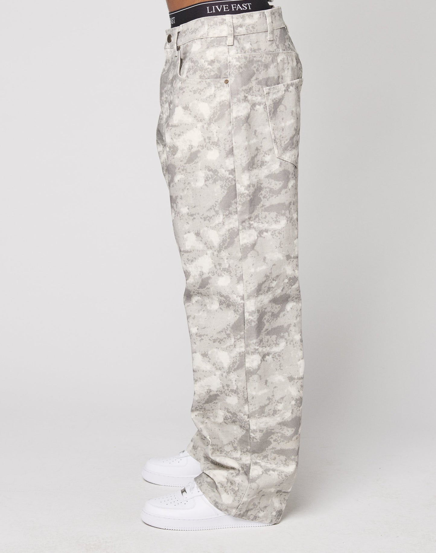A person stands sideways in LIVE FAST DIE YOUNG’s Live Fast Canvas Pants—light gray camo with a baggy fit and wide straight leg—paired with white sneakers and a black LIVE FAST waistband, set against a plain white background.